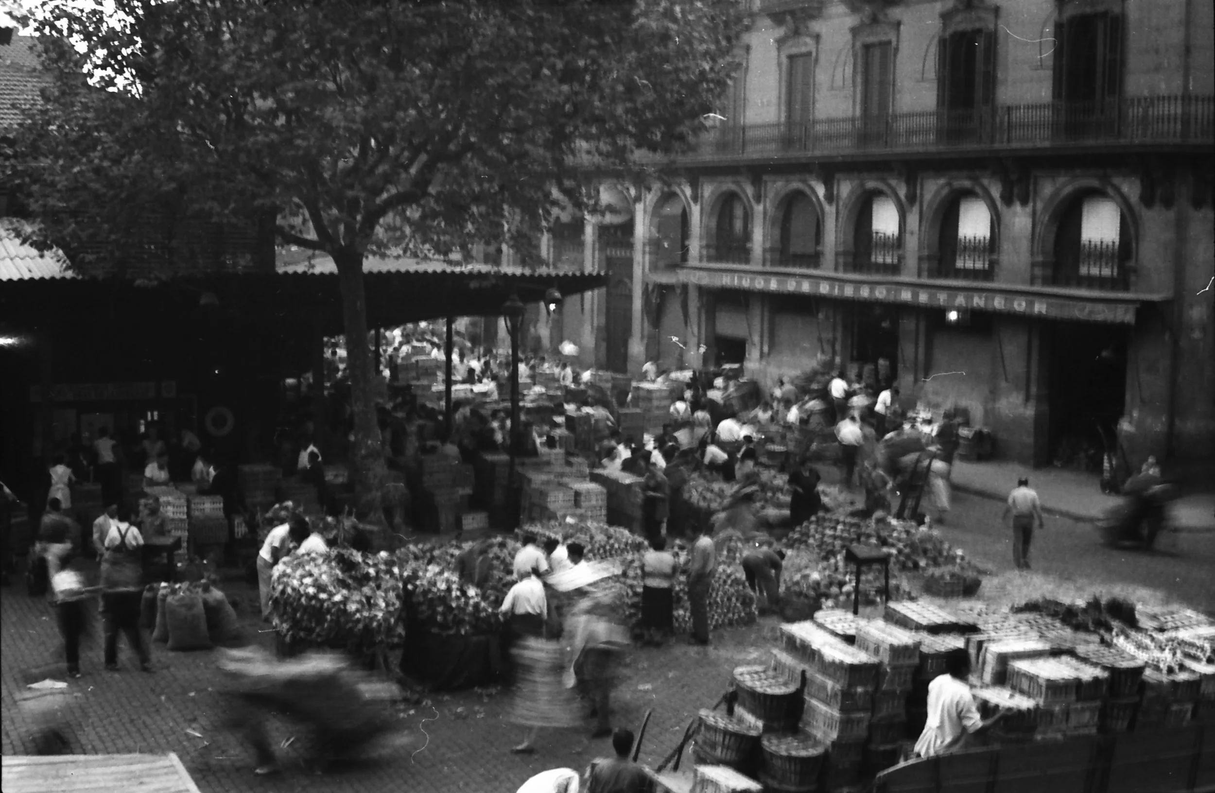 A busy outdoor market with numerous people shopping and selling goods, and a large building in the background with arched windows and iron balconies.