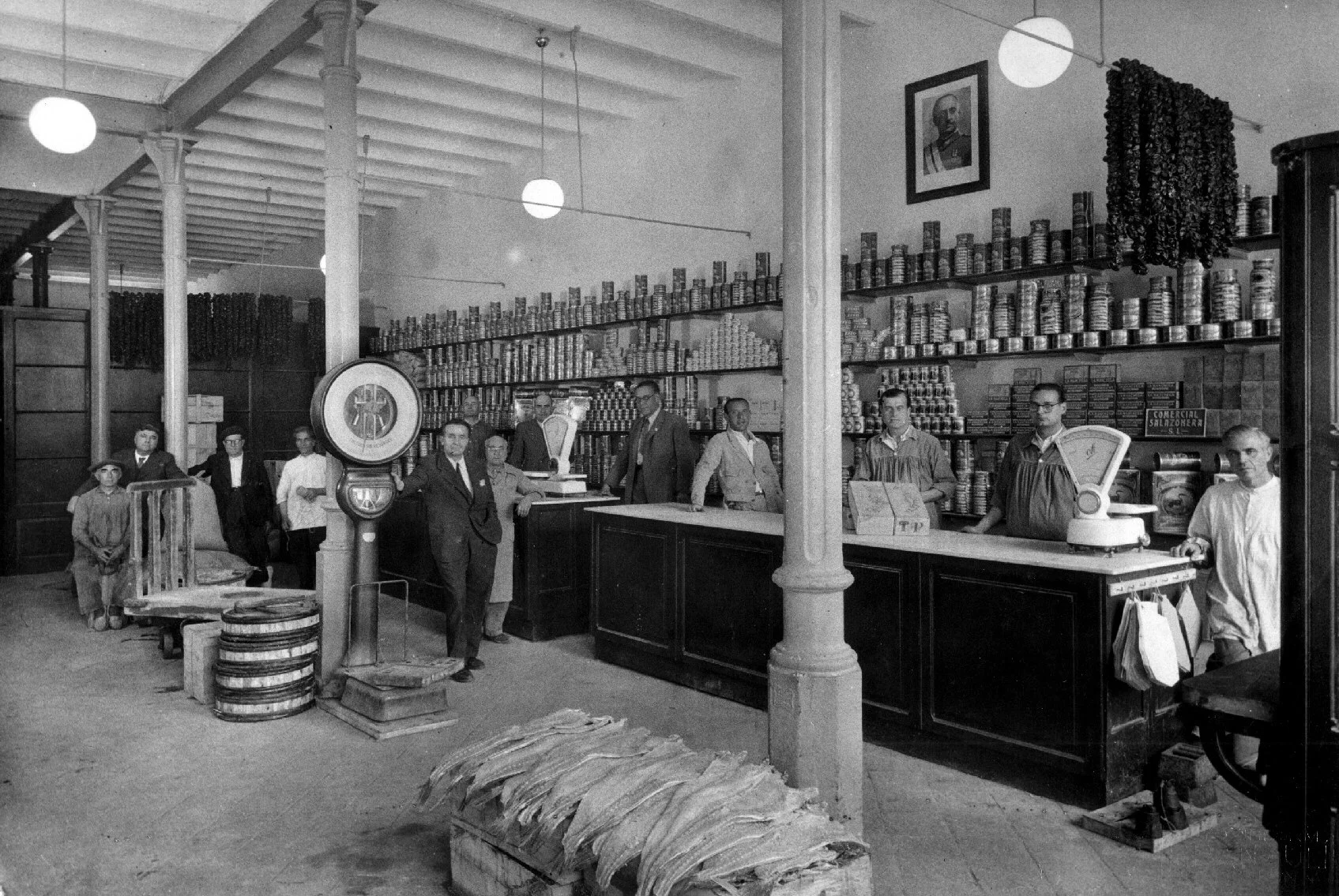 Black and white photo of a grocery store counter with multiple workers behind it, shelves filled with cans, a scale, and a vintage scale in the foreground. Several people stand near the counter, and a portrait of a man is hanging on the wall above.