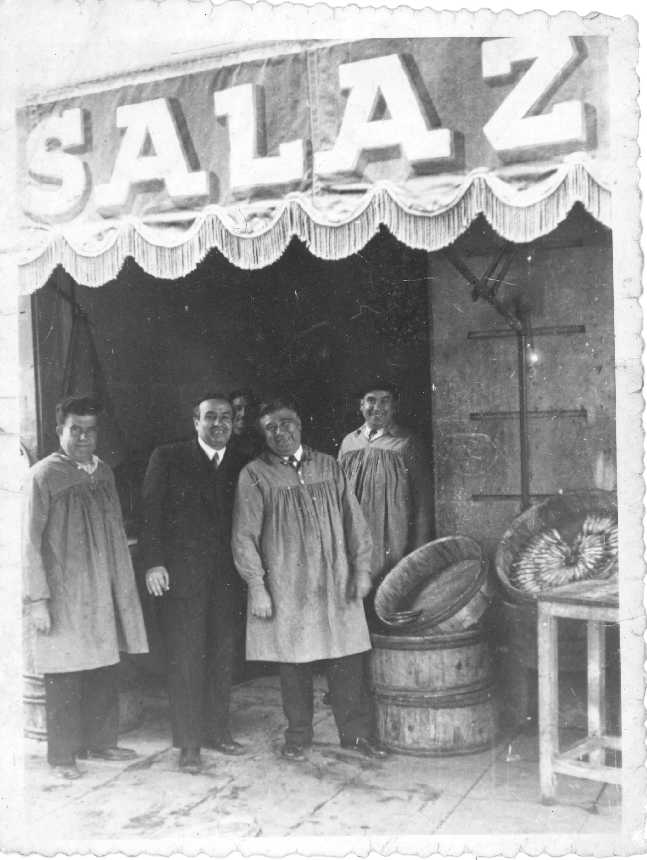 Black and white photo of five men standing in front of a carnival carnival game booth with a sign reading "Salaz". The booth is decorated with fringe and has large candy display jars on the front table.