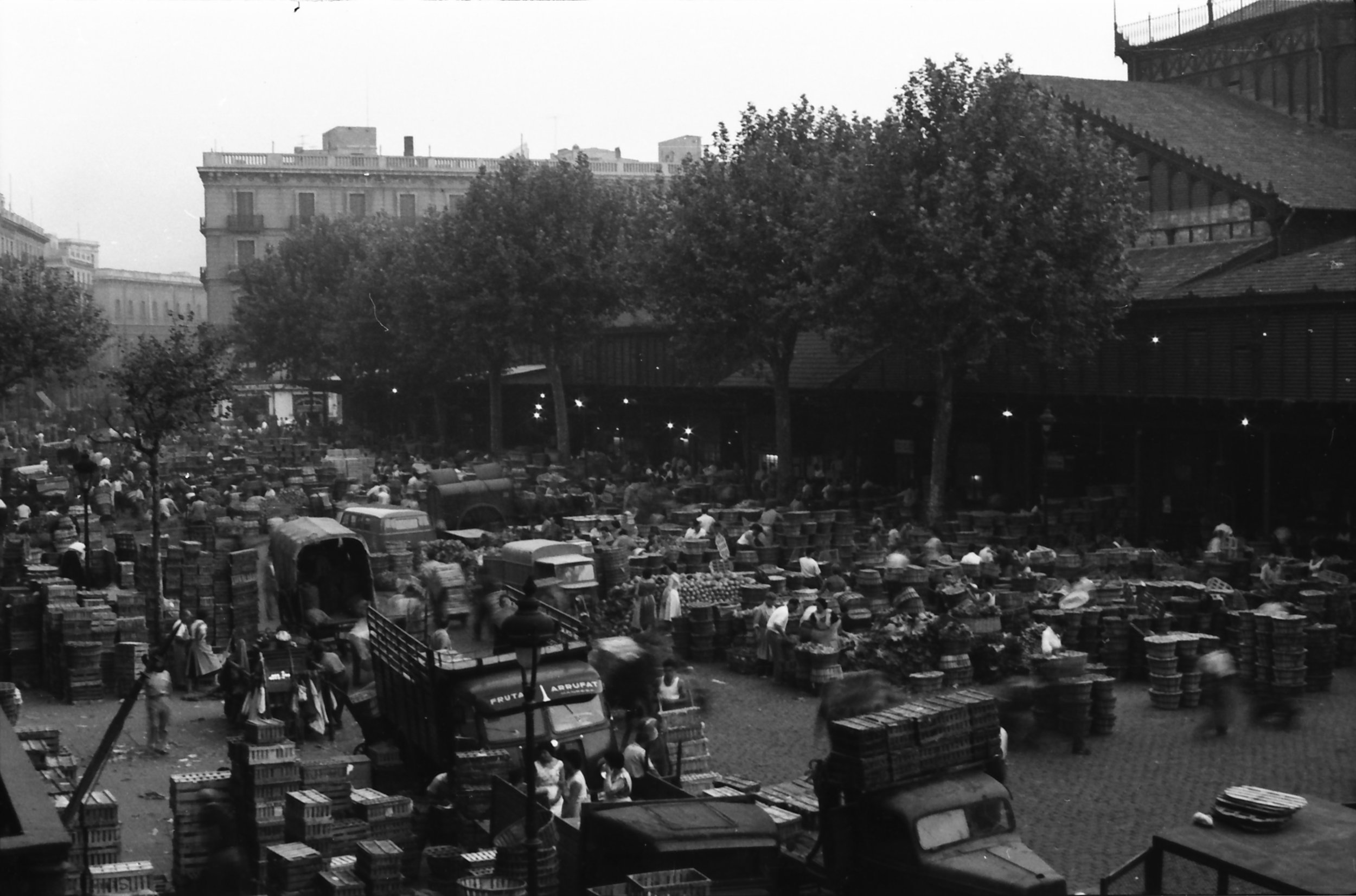 Black and white photograph of an outdoor market with many people, stalls, and crates filled with fruits and vegetables, situated in an urban area surrounded by buildings and trees.
