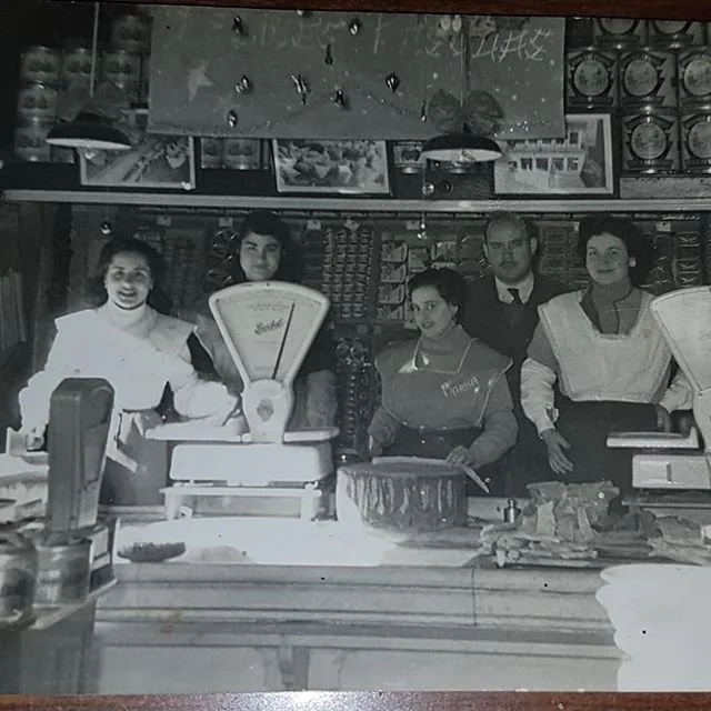 Black and white photo of five people working at a bakery or shop counter, with scales and baked goods on the counter, and shelves stocked with products behind them.