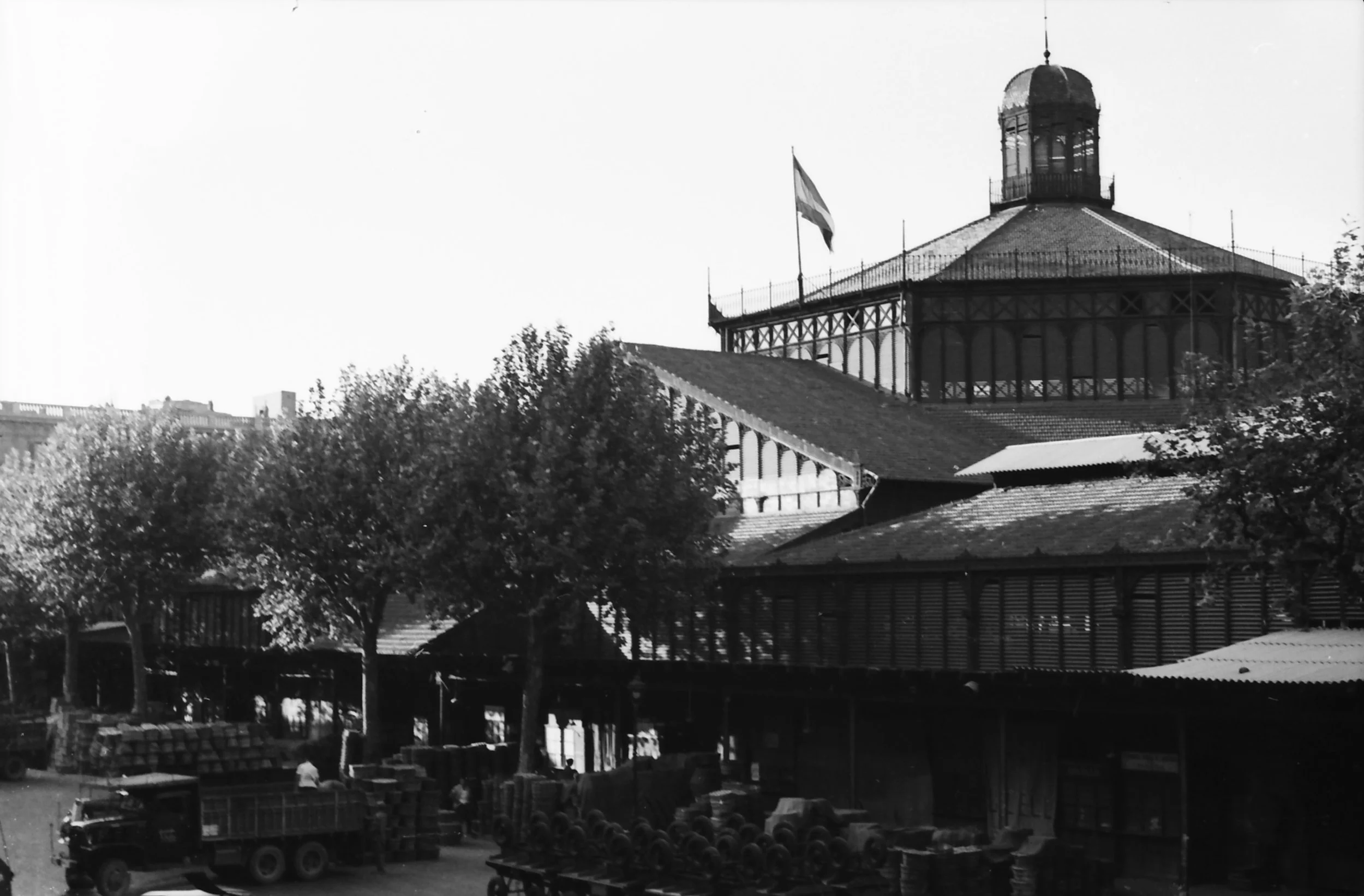 Black and white photo of a historic building with a dome and a flag, trees in the foreground, and a market scene below with trucks and people.