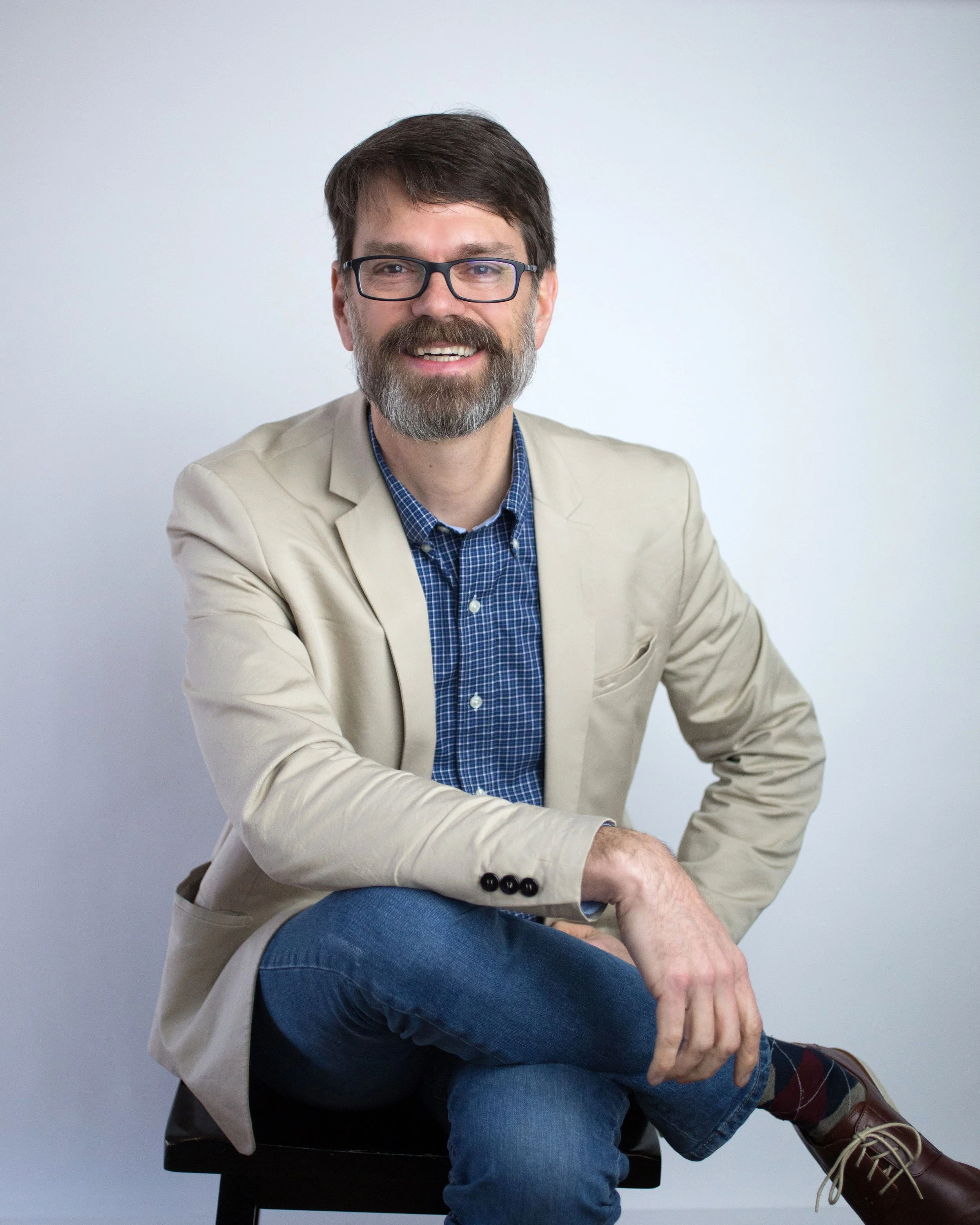 Prof C wearing a beige blazer and blue checkered shirt, sitting on a black chair.