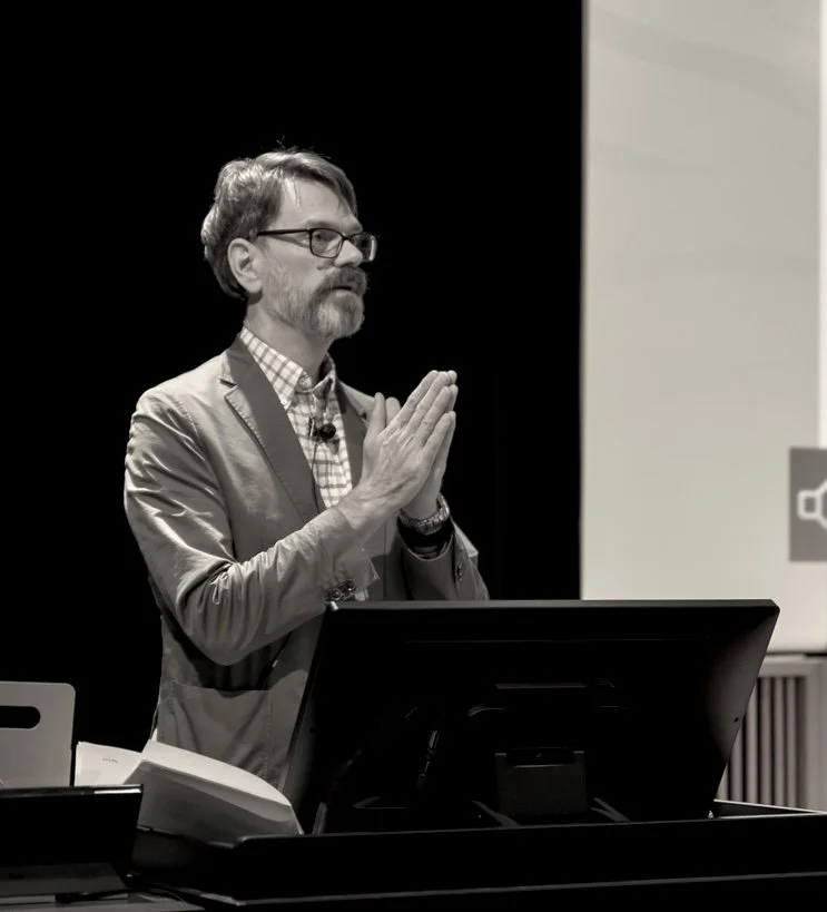 Prof C standing behind a desk with a computer monitor, possibly giving a presentation or lecture, with a dark background and a light-colored wall to the side.