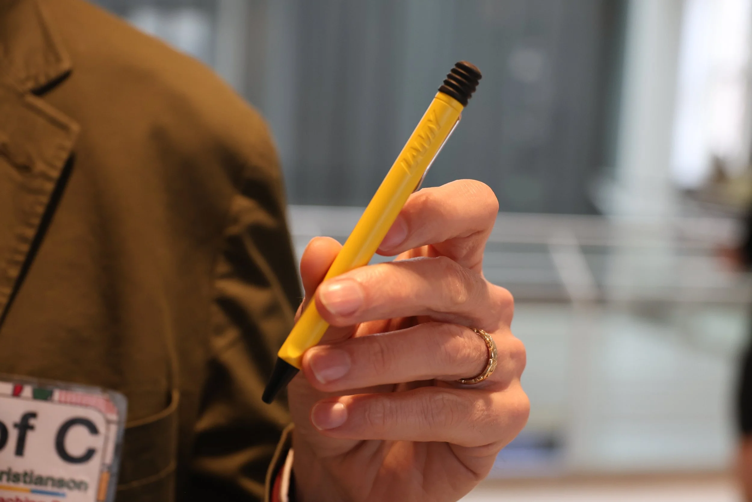 Close-up of Prof C's  hand holding a yellow pencil with a black eraser, wearing a gold ring on the ring finger, in an indoor setting.
