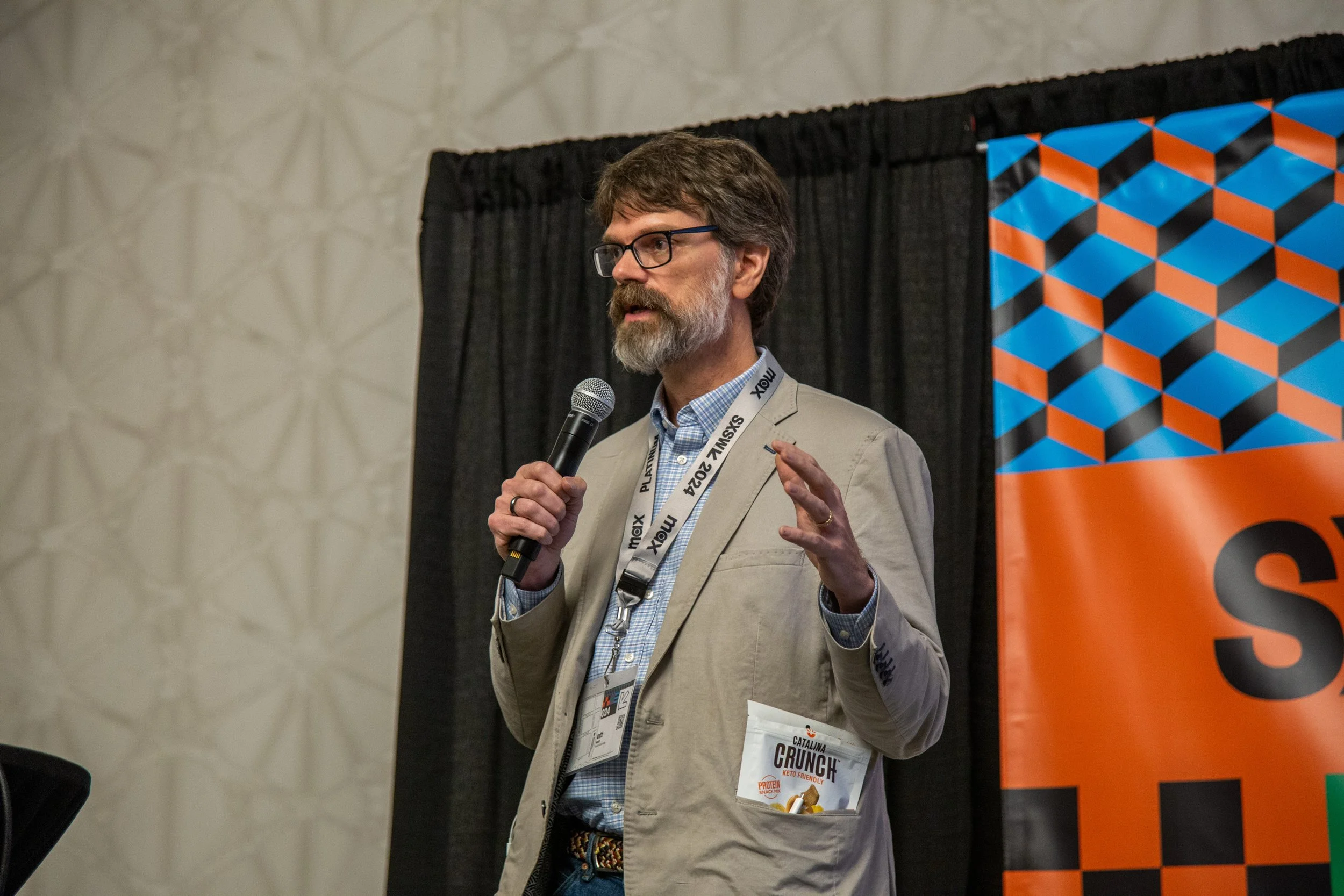 Prof C speaking into a microphone at the SxSW conference, with a black curtain and colorful geometric banner in the background.