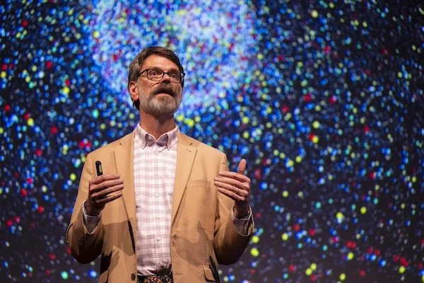 Prof C giving a presentation on stage with a colorful, abstract background of illuminated dots.