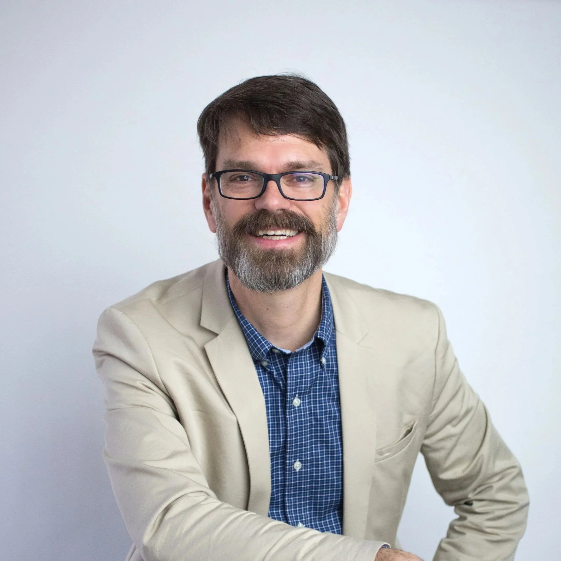 Prof C wearing a beige blazer and blue checkered shirt, sitting against a plain white background.