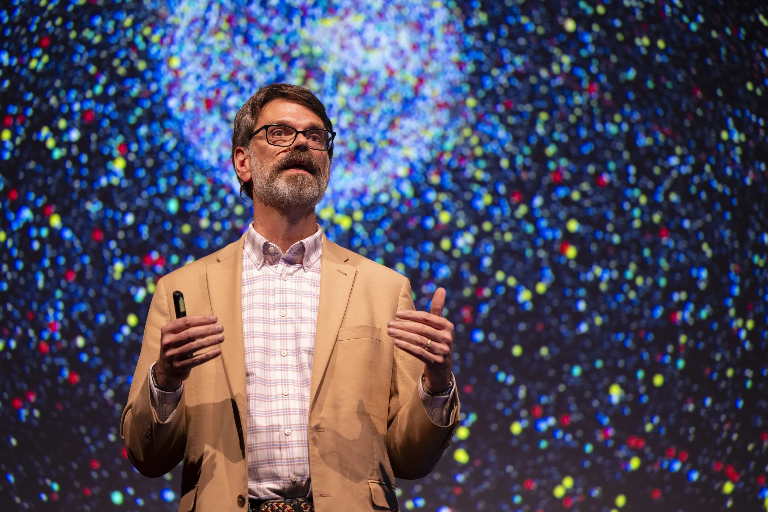Prof C wearing a beige blazer and checked shirt giving speech on stage with a colorful, starfield background.