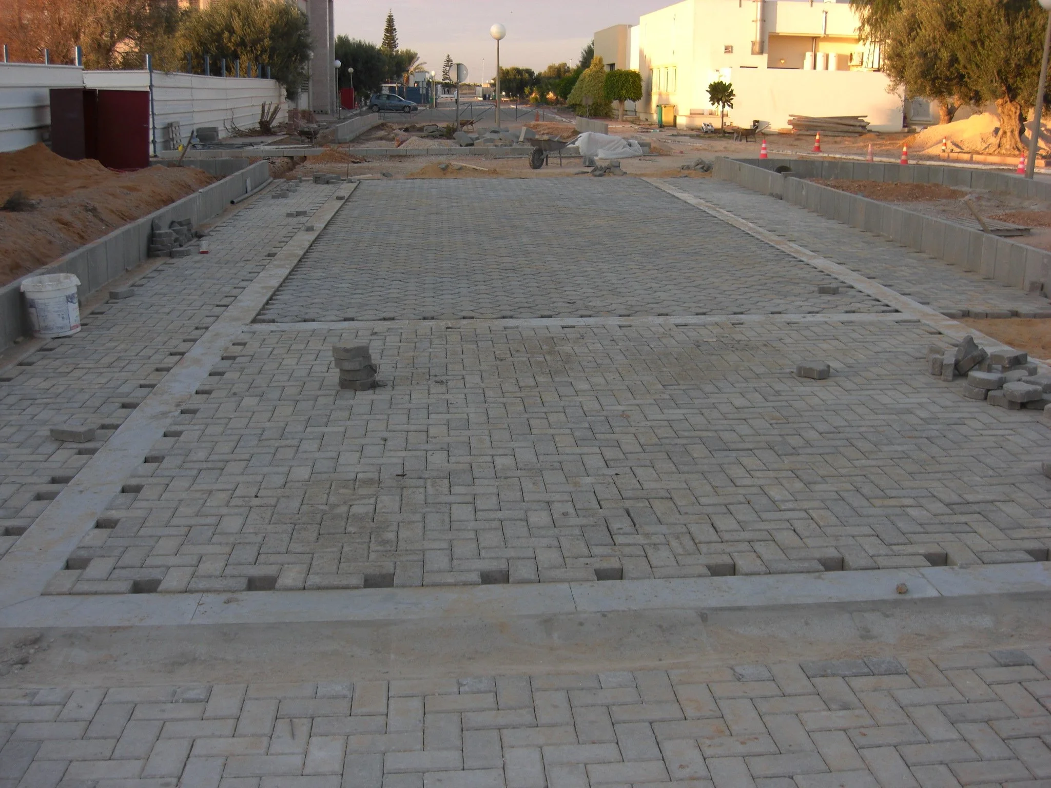 Construction site where bricks are being laid for a new sidewalk or road. Piles of bricks and construction materials are visible, with some partially laid and others waiting to be installed. In the background, there are trees, buildings, and traffic cones indicating ongoing construction work.