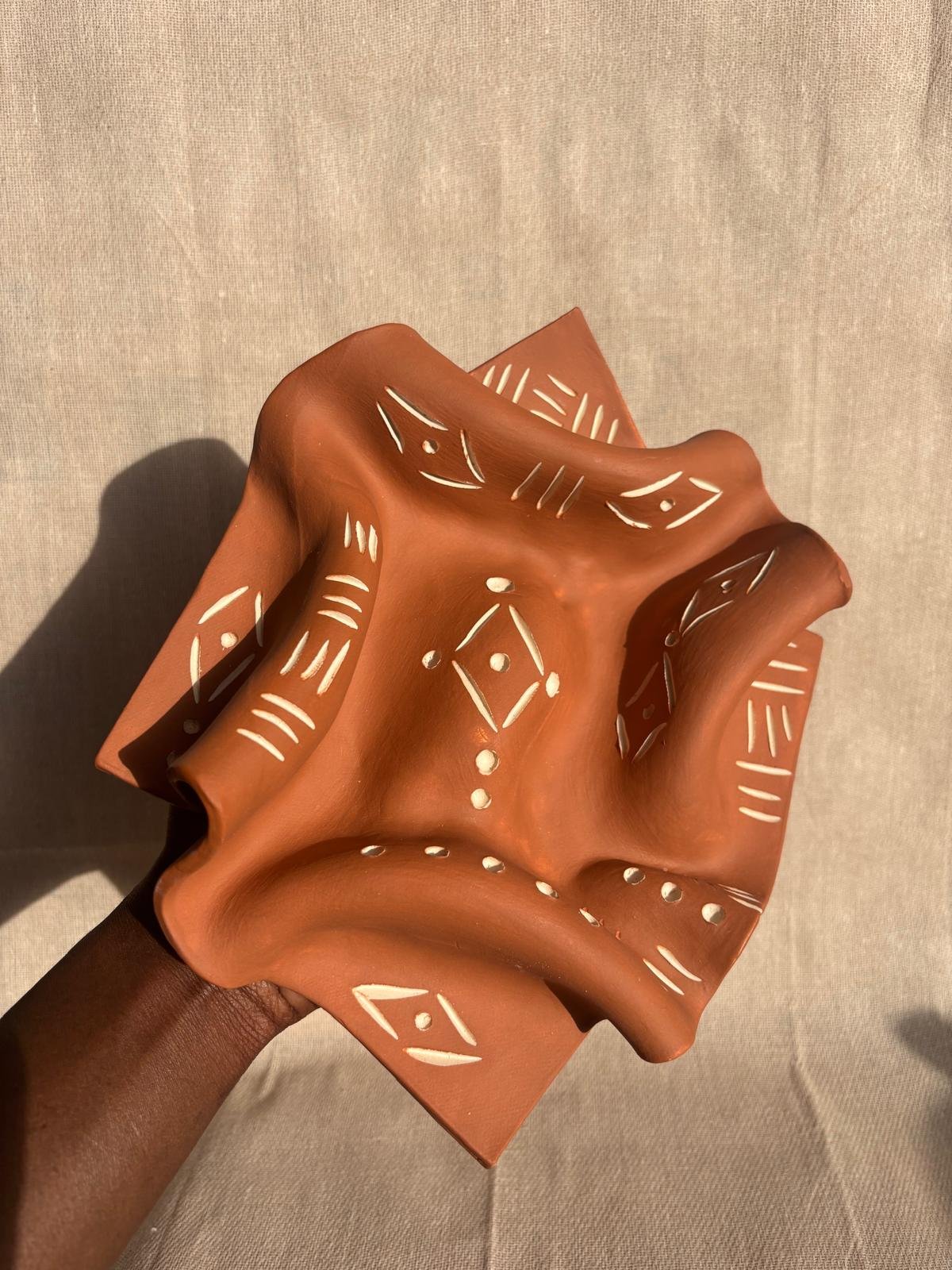 A terracotta-colored ceramic sculpture with abstract, flowing shapes and white geometric patterns painted on its surface, held against a light-colored background.