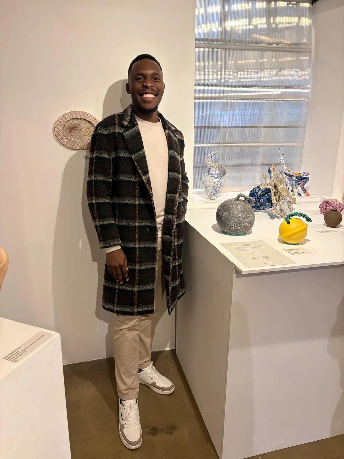 A man smiling and standing near a display of art sculptures in a gallery. He is wearing a plaid coat, white shirt, beige pants, and white sneakers.