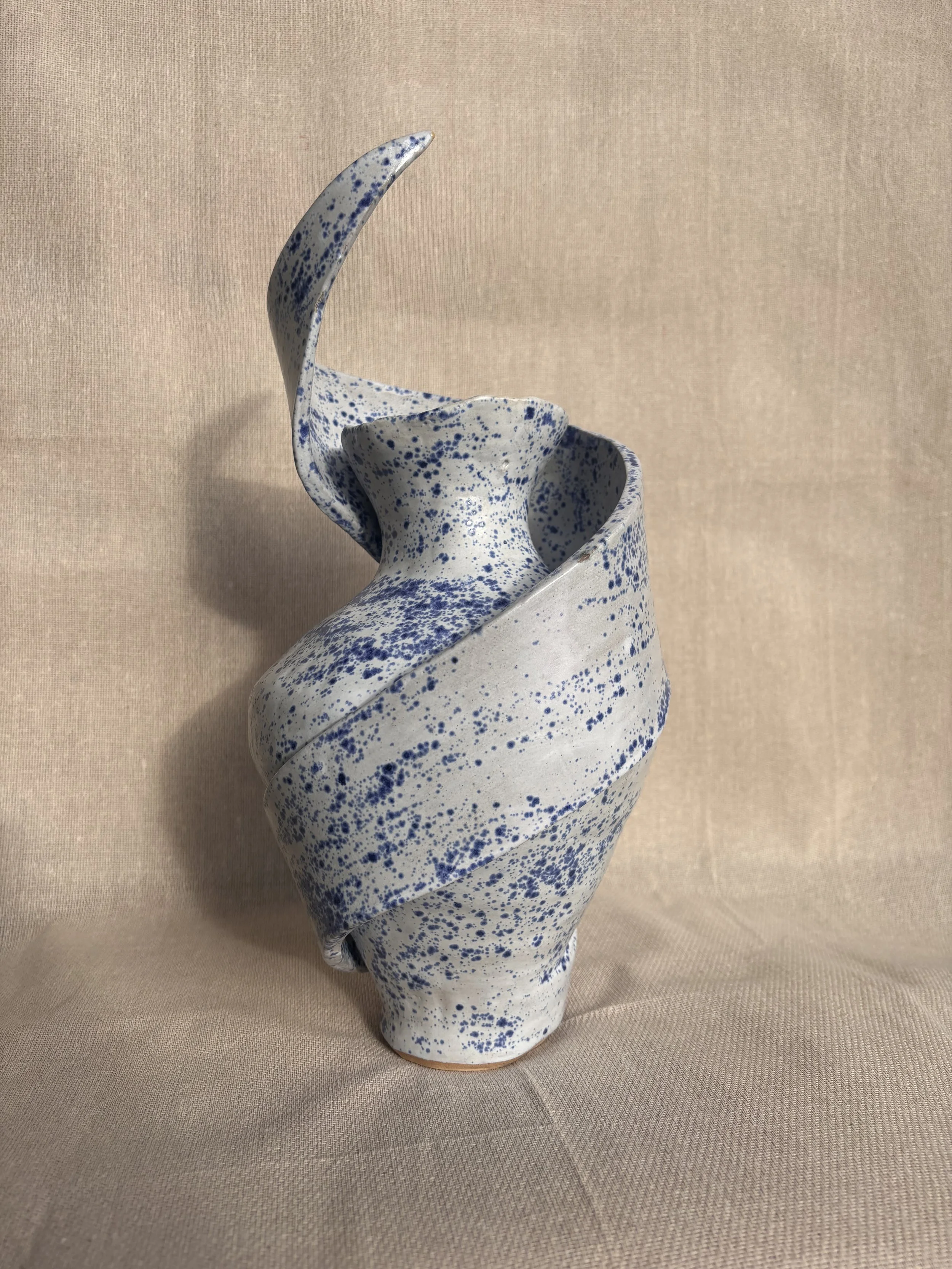 A tall ceramic vase with a narrow neck and a wider base, decorated with blue speckles on a white background, placed on a fabric surface with a neutral background.