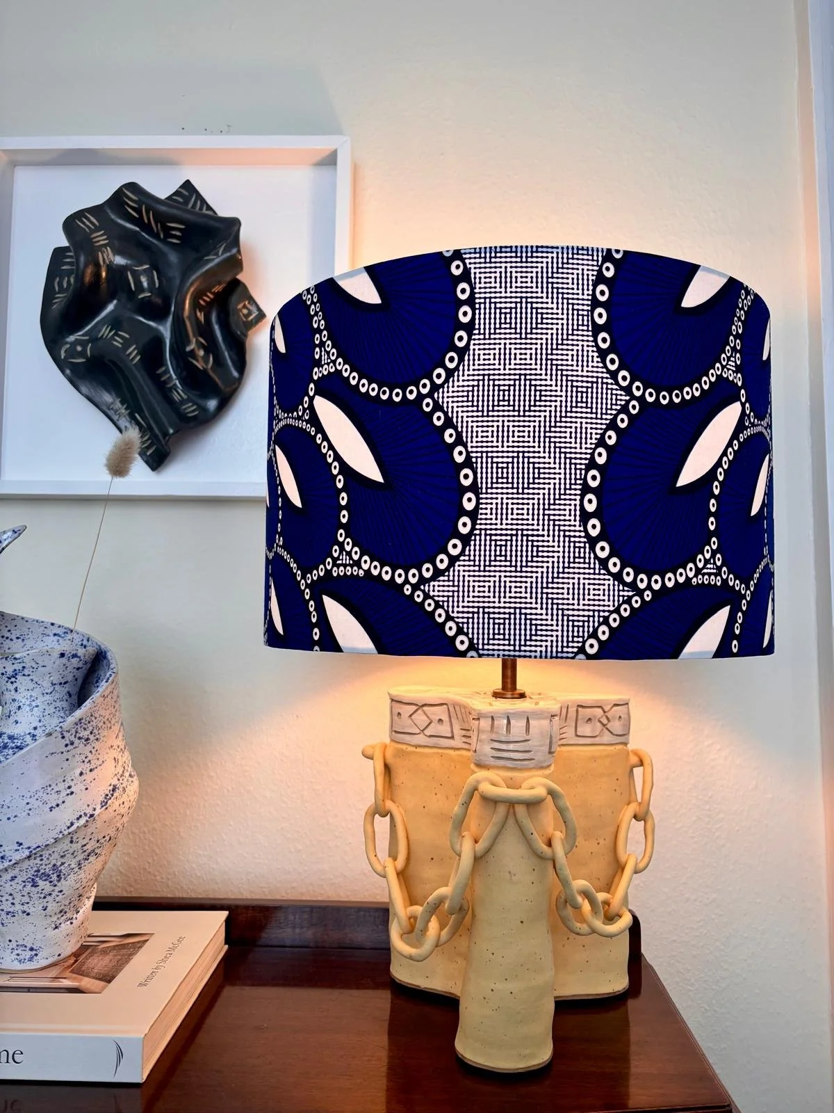 A decorative table lamp with a patterned shade featuring blue and white geometric design, set on a ceramic base with chain links. To the left, a large ceramic vase with blue speckles, partially visible, and a black sculpture of a crumpled shape mount