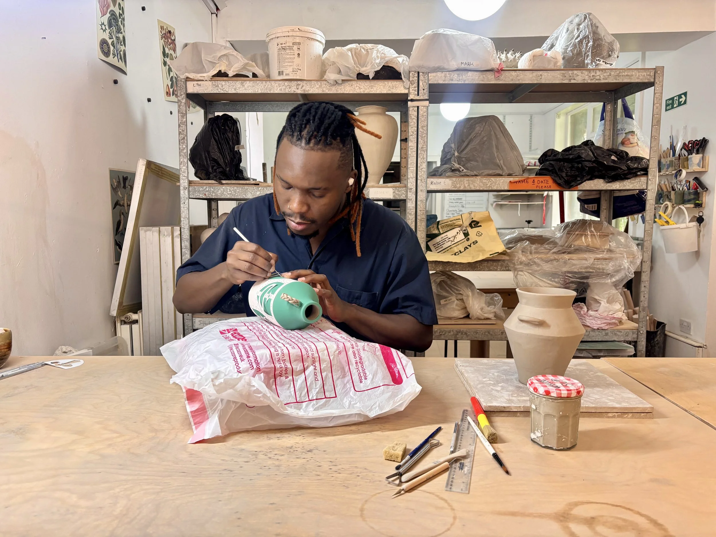 A person with dreadlocks working on a pottery project in a ceramics studio, surrounded by pottery tools, supplies, and unfinished ceramic pieces on shelves.