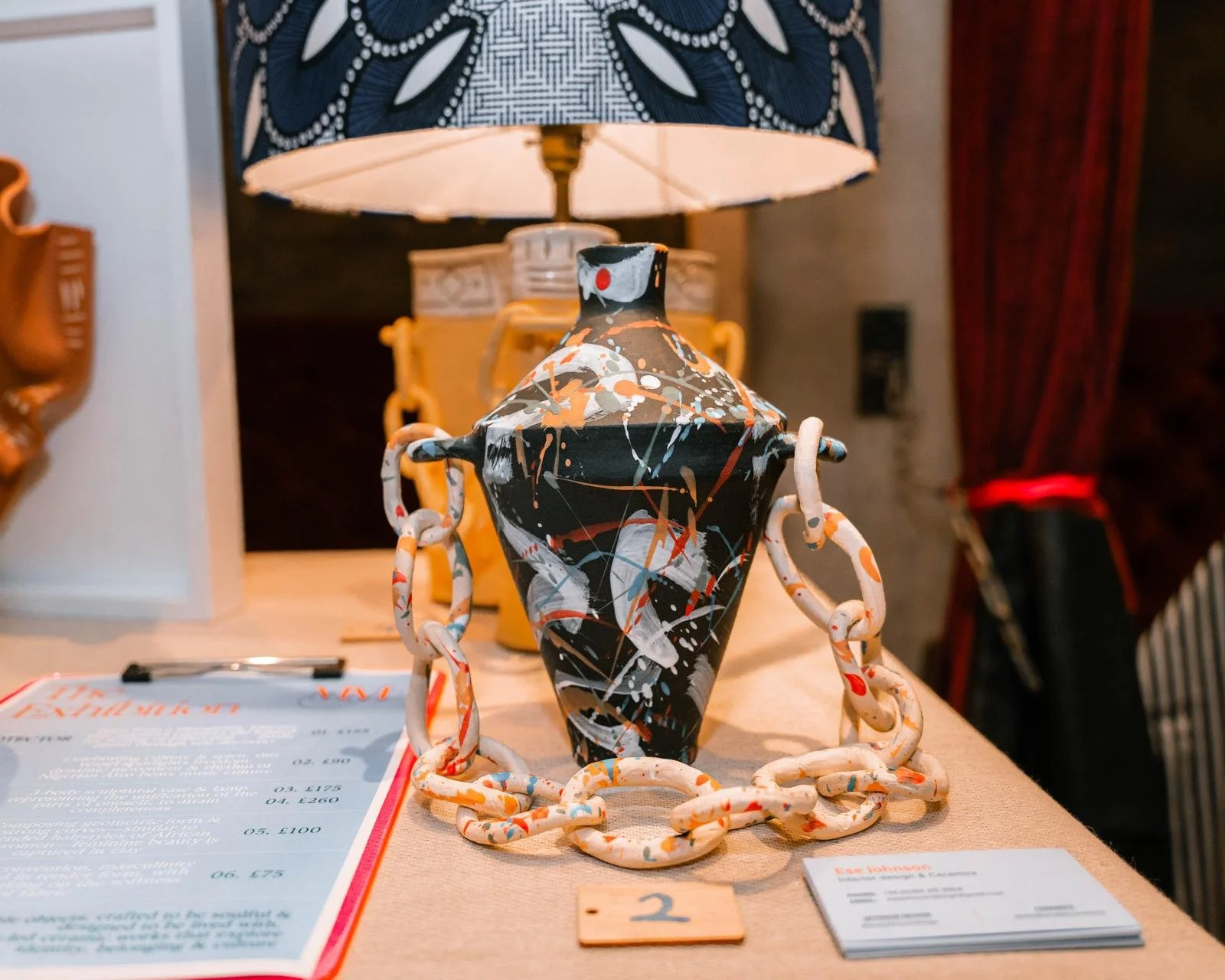 A decorative ceramic vase with an abstract splatter paint design in black, white, orange, and gray, surrounded by a chain-like sculpture, displayed on a table in an art gallery or museum.