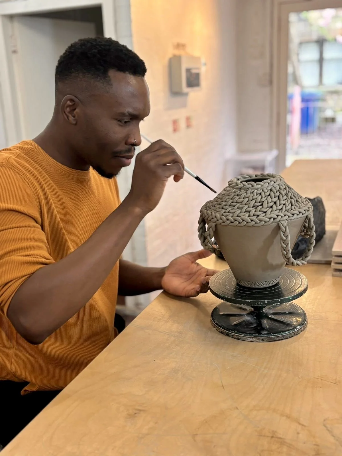 A man in an orange shirt is painting a large ceramic vessel with detailed, rope-like handles. The vessel is on a rotating turntable on a wooden table in a bright, airy room.