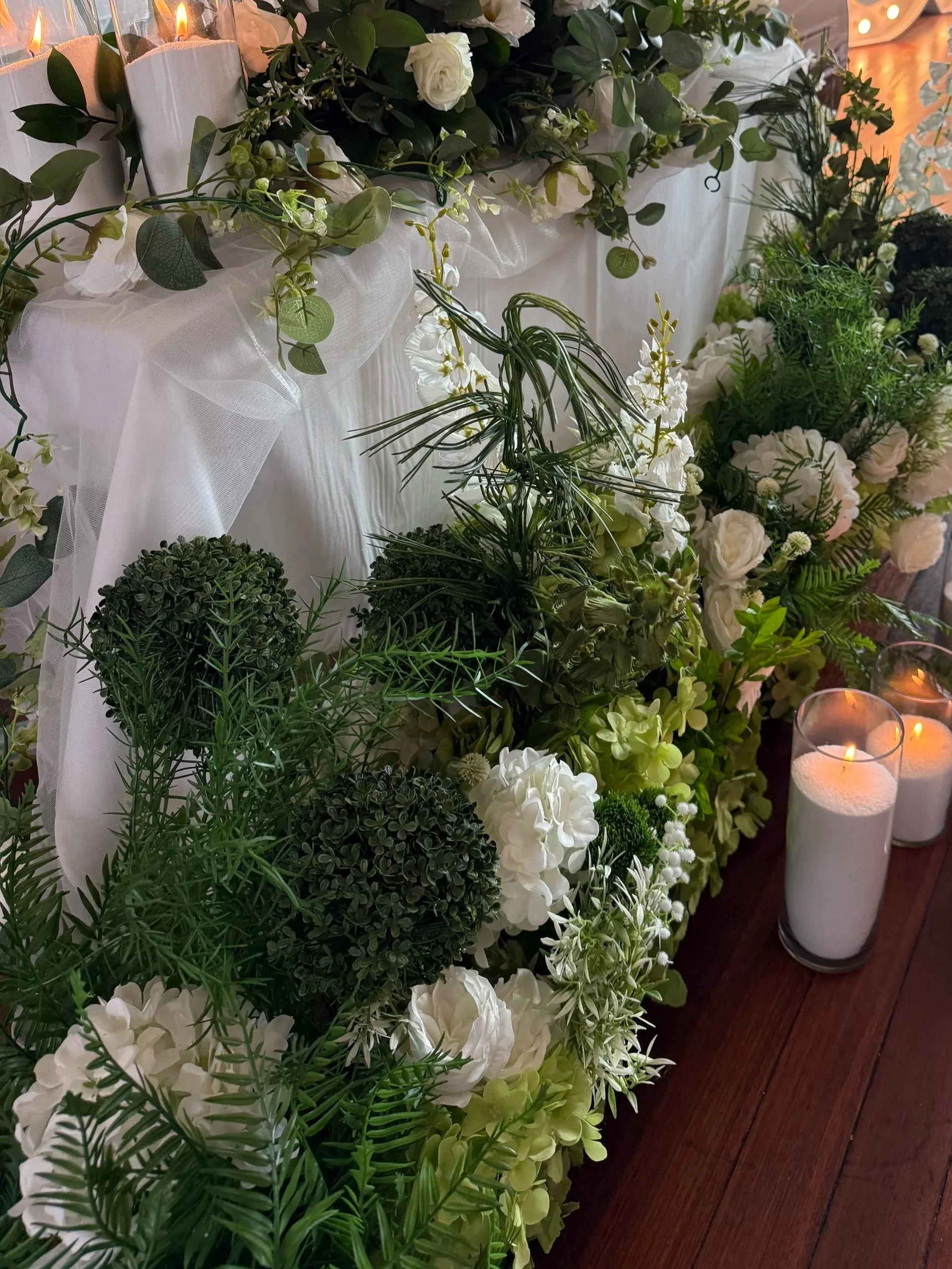 A floral arrangement with white and green flowers and foliage, illuminated by candles in glass holders, on a dark wooden surface, possibly for a wedding or special event.