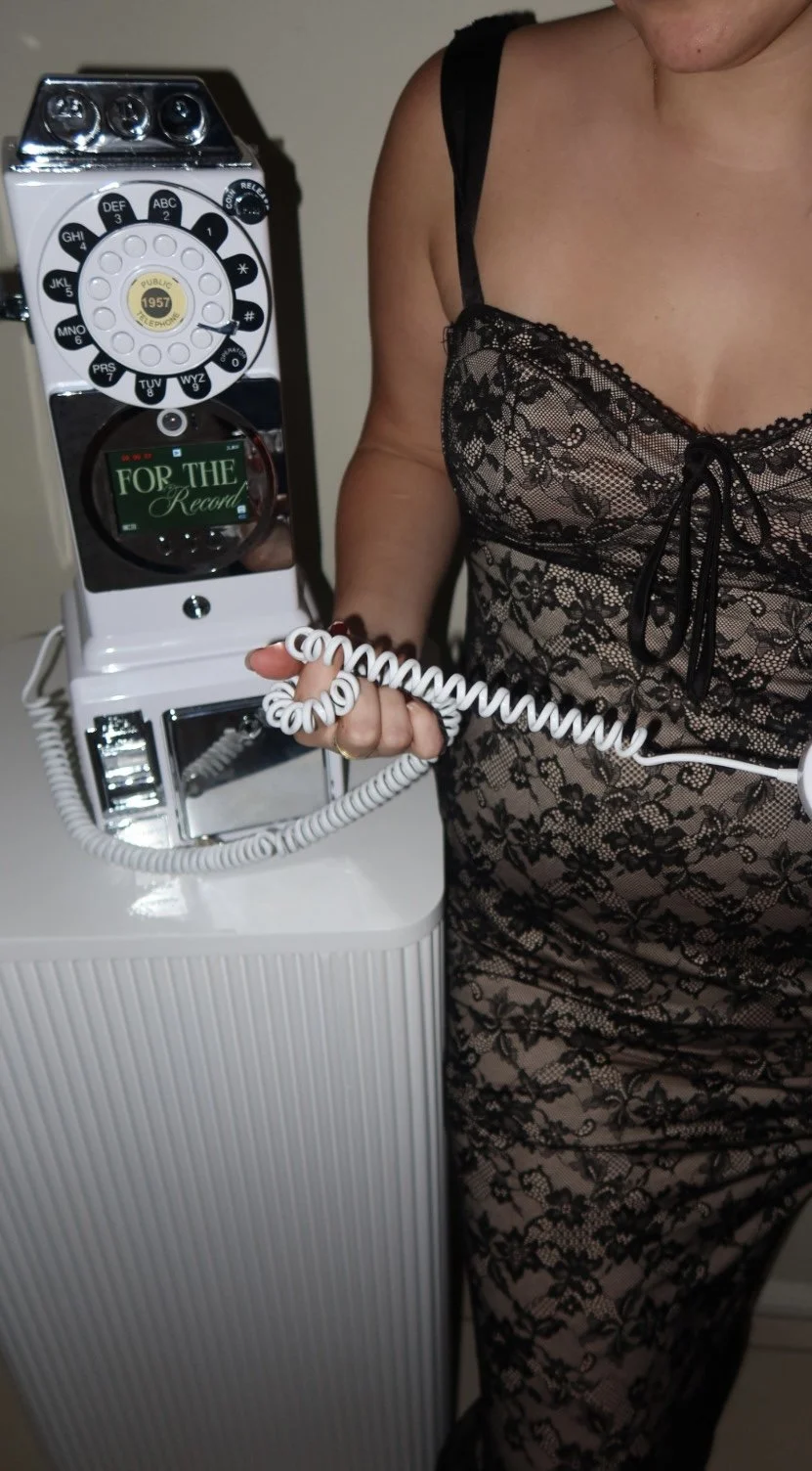 Person in a black lace dress holding a vintage rotary telephone with a display that reads "FOR THE Record."