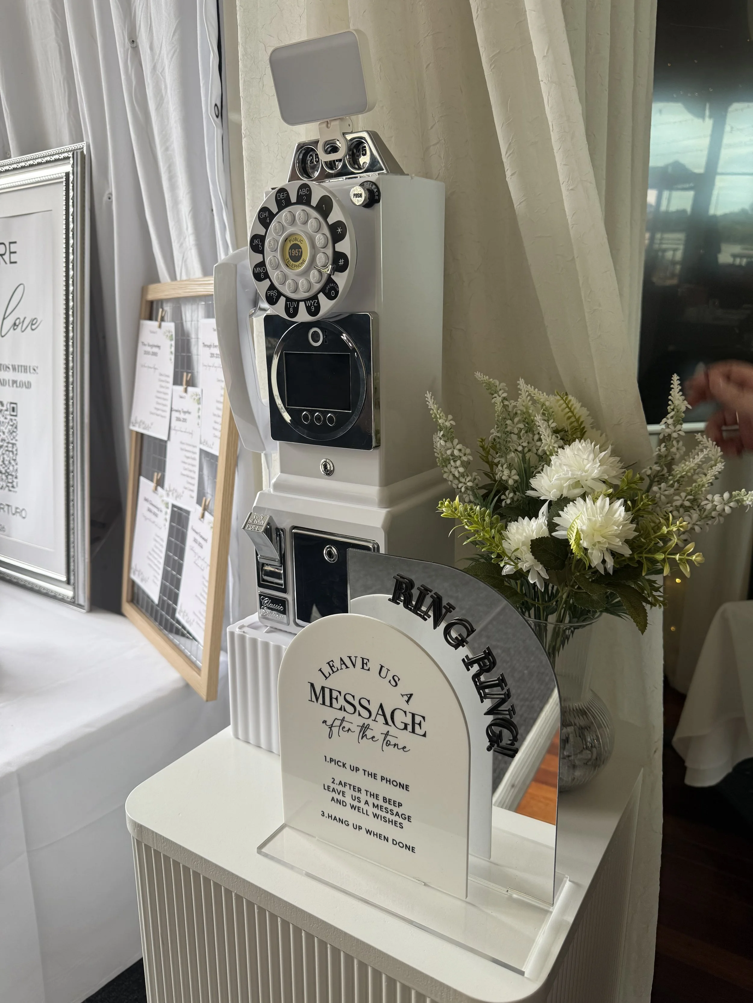 A white vintage telephone on a table with a sign explaining a message leaving procedure, next to a white vase with white flowers, in a setting with light-colored curtains and a window.