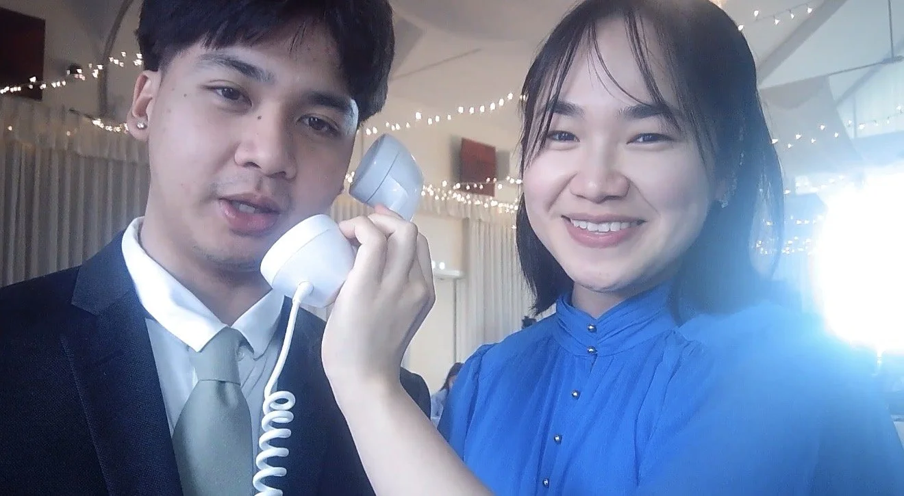A young man in a suit and tie speaking on a corded phone, smiling, while a young woman in a blue dress holds the phone to his ear, smiling at the camera, with string lights in the background.