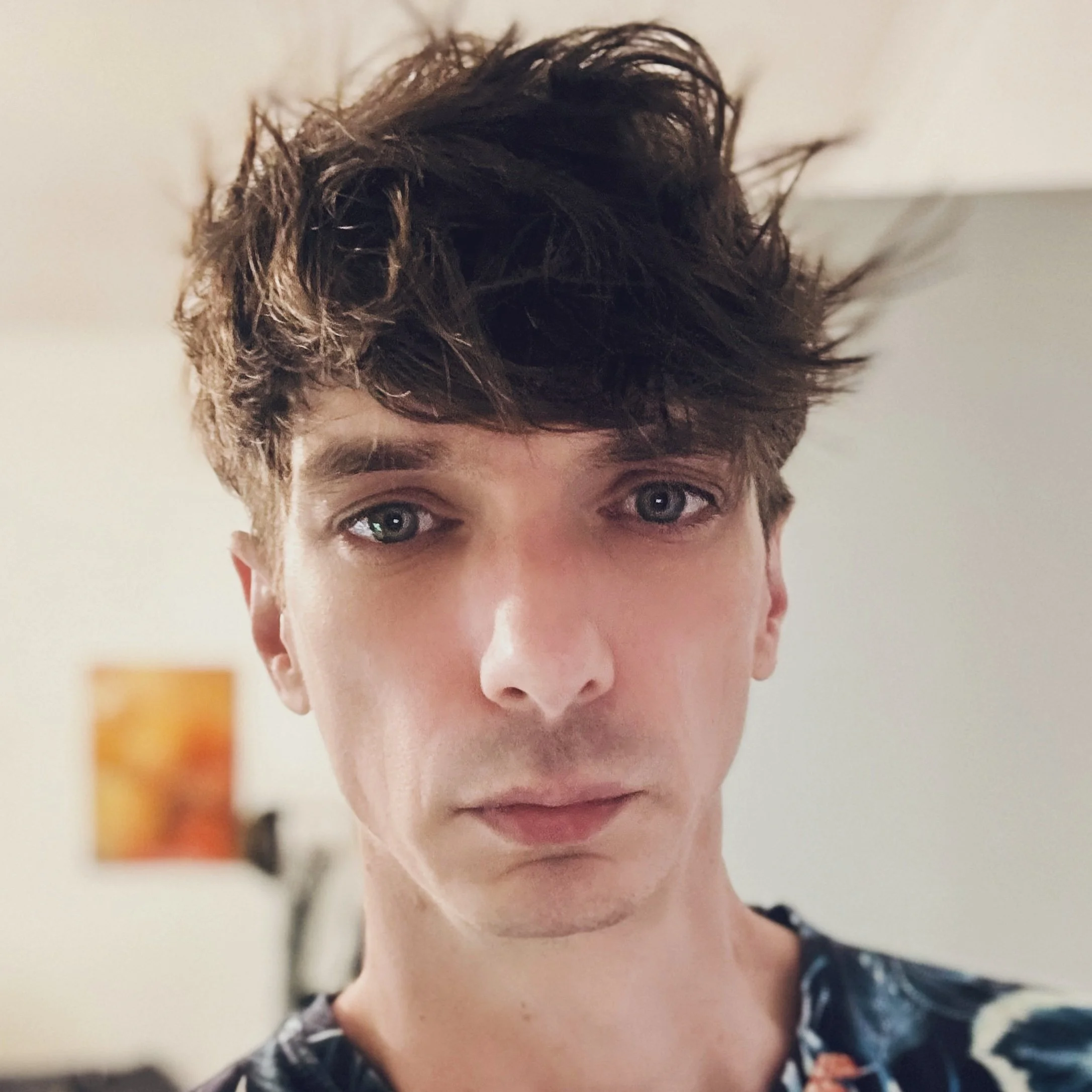 Close-up of a young person's face with short, curly brown hair and blue eyes, wearing a denim jacket with a patterned shirt underneath.
