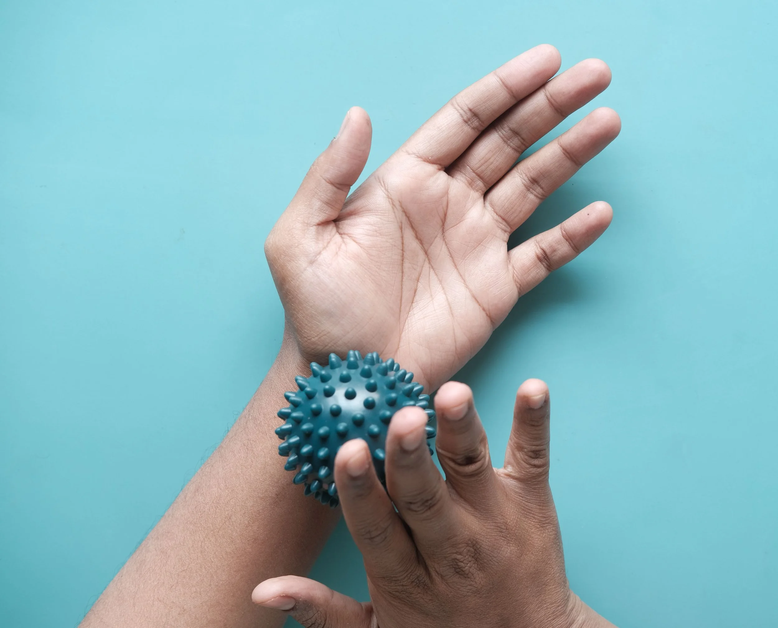 Close-up of a person's hand with a blue spiky rubber massage ball on the wrist, against a light blue background.