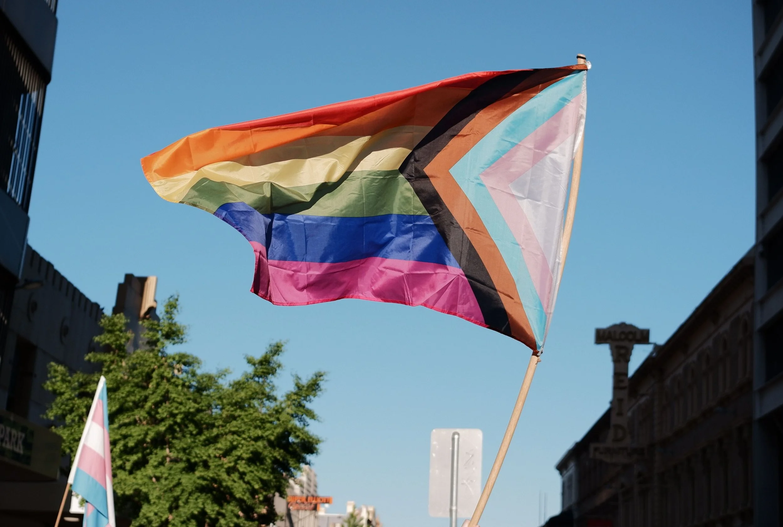 A rainbow flag with stripes of orange, yellow, green, blue, pink, and black, waving in the wind against a clear blue sky in an urban area.