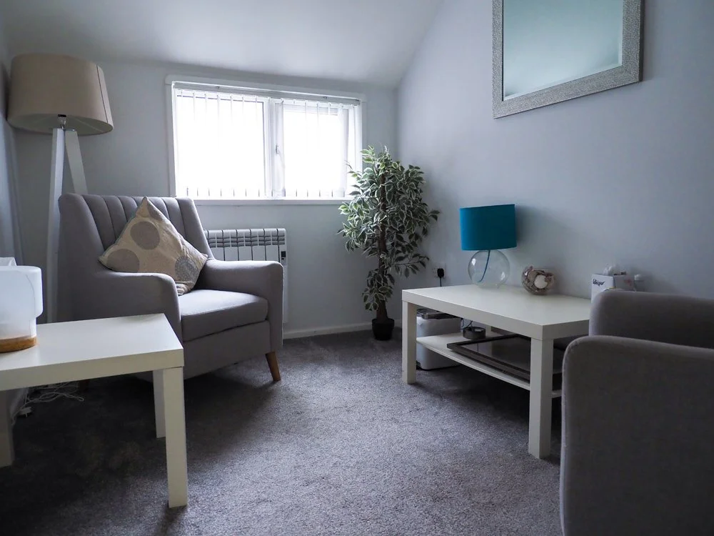 A cozy counselling room with a gray armchair, a white side table, a white coffee table, a large potted plant, a window with blinds, a teal table lamp, and a mirror on the wall.