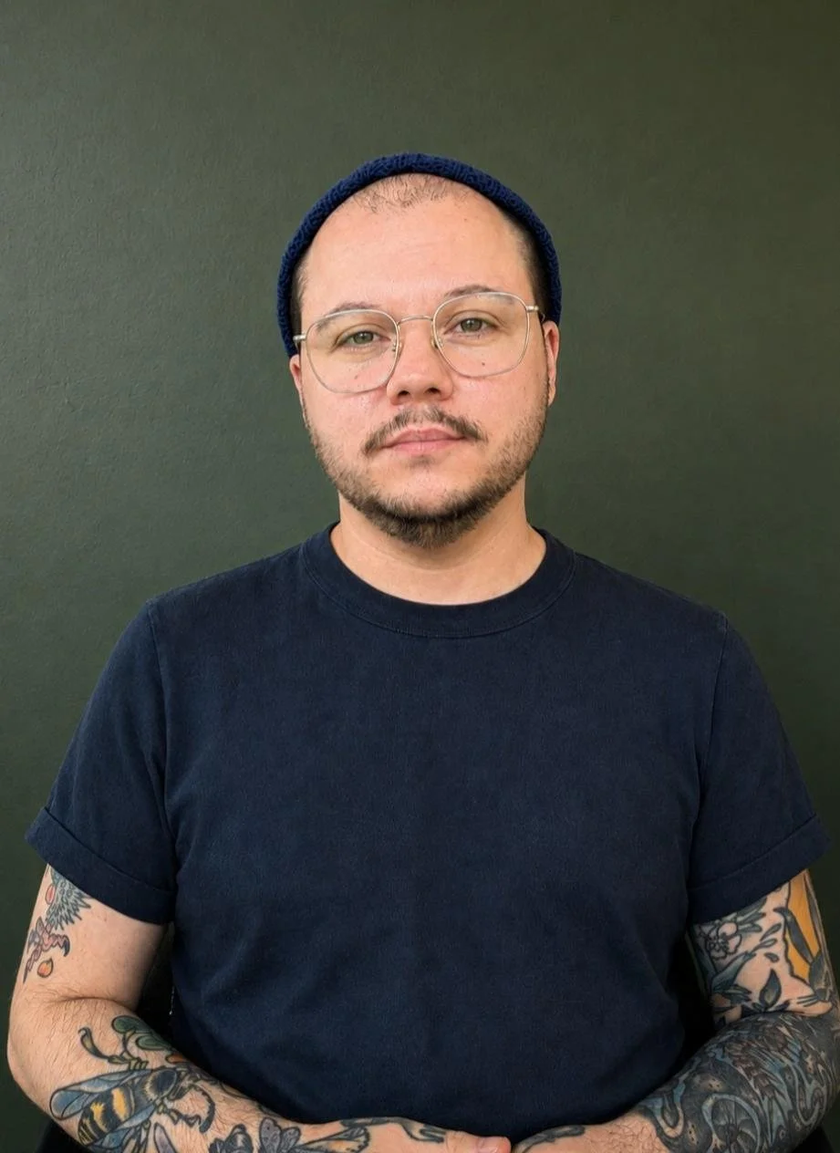 A man with glasses, a beard, and tattoos on his arms wearing a dark t-shirt and a navy beanie against a plain dark background.