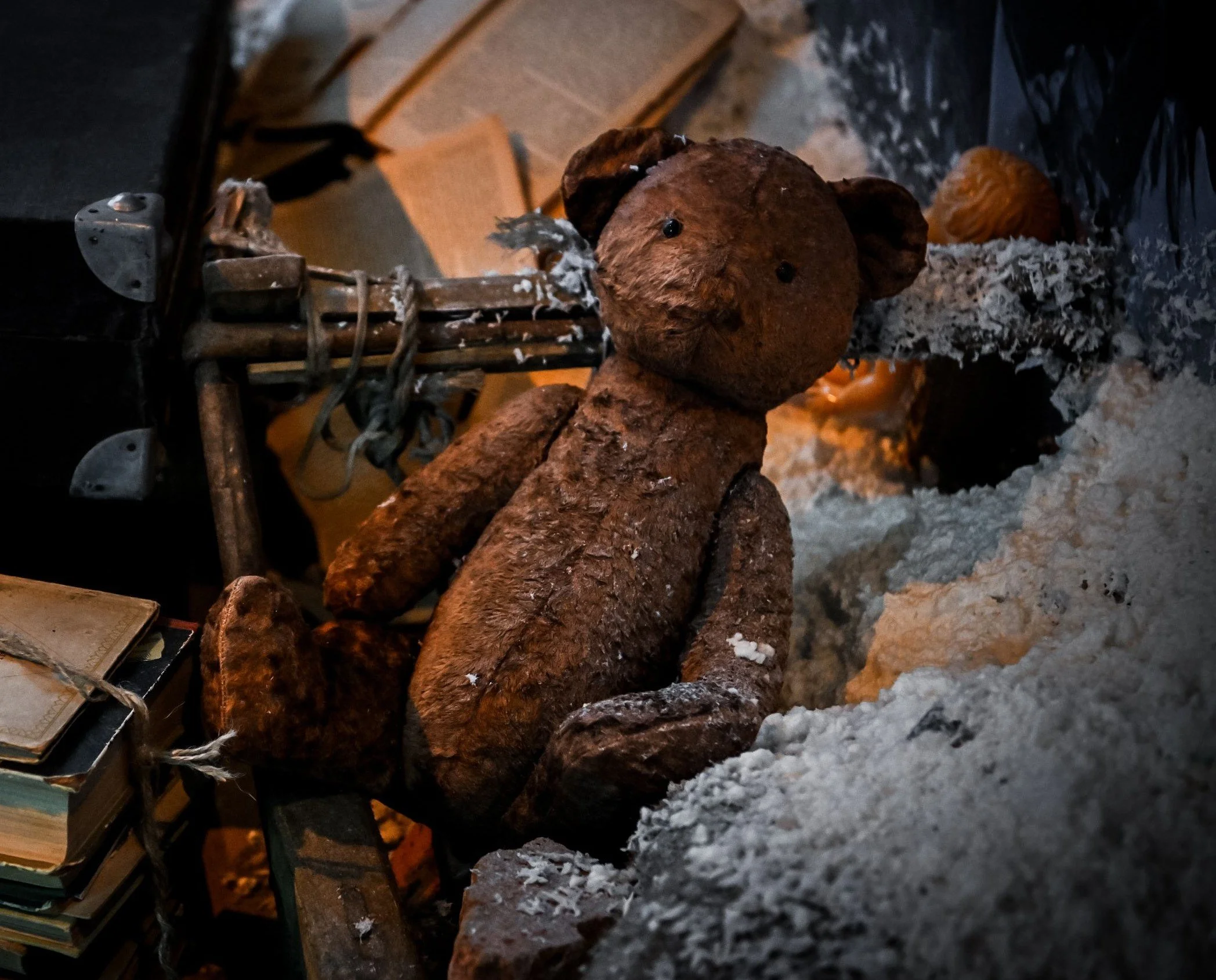 A close-up of a small teddy bear figurine made of brown, textured material with black eyes, sitting on a surface surrounded by scattered gray and white shredded paper or ash, with a metal object and old books nearby.