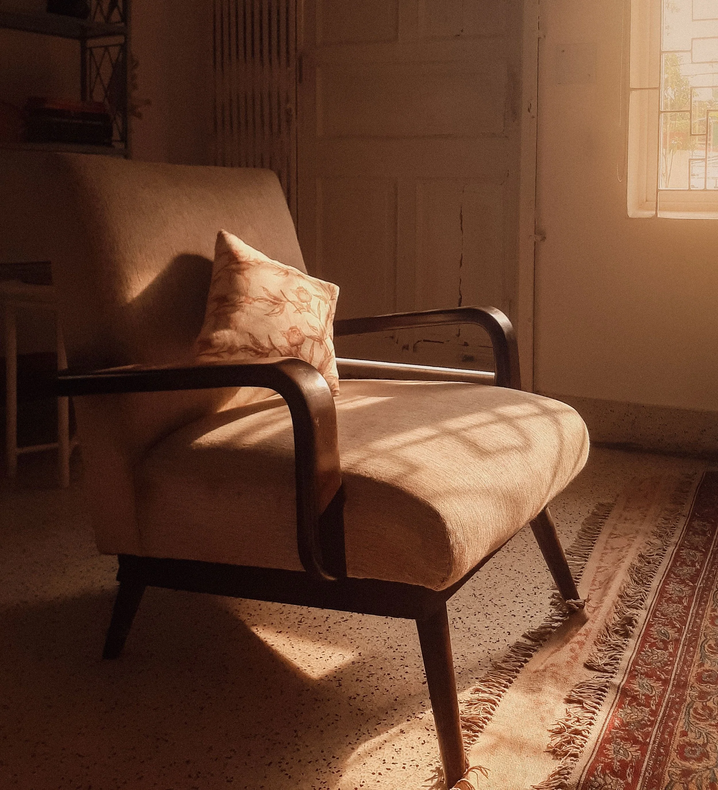 A vintage-style room with a wooden armchair, a floral cushion, a patterned rug, and sunlight coming through a window.