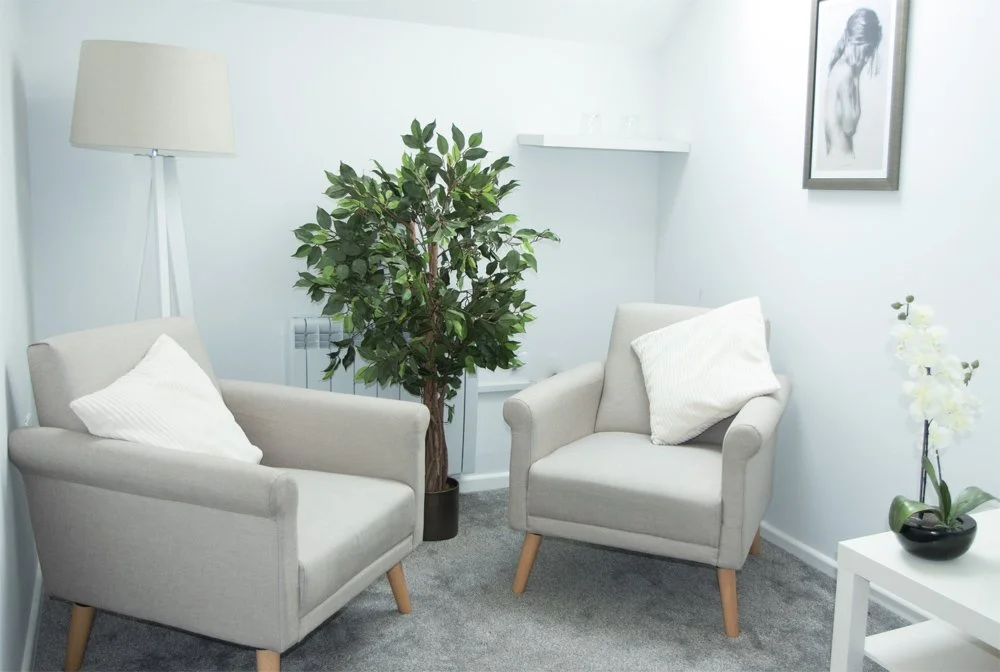 A cozy seating area with two light beige armchairs with white pillows, a tall floor lamp, a large houseplant, a framed black and white art photo on the wall, and a white side table with a potted orchid on it.
