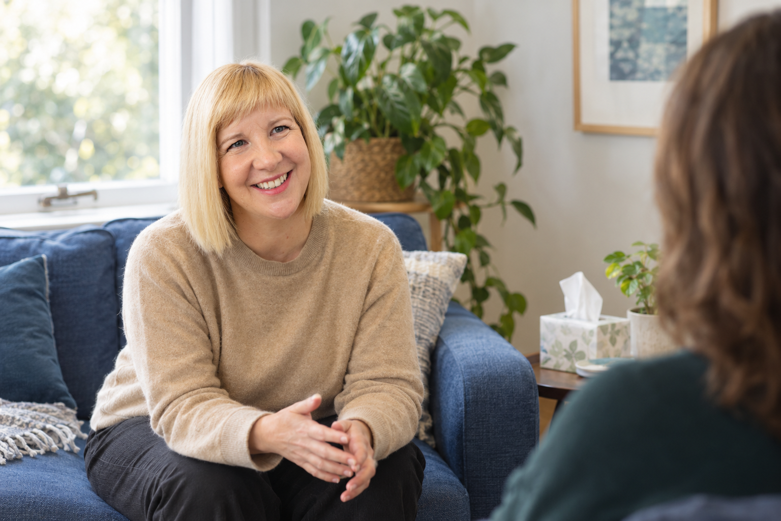 Sarah Black sitting on a sofa during a conversation with a psychotherapy client in a cozy therapy room with houseplants and window.