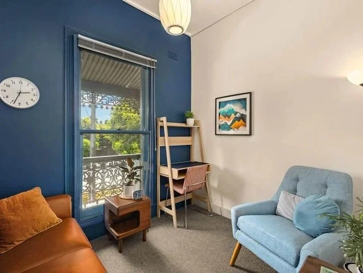 room with blue and white walls, a window with decorative ironwork, a clock, a small wooden table with a plant and a telephone, a brown sofa with an orange pillow, a light blue armchair with a gray pillow, a framed mountain landscape painting, a pine tree, and a wooden desk with a pink chair.