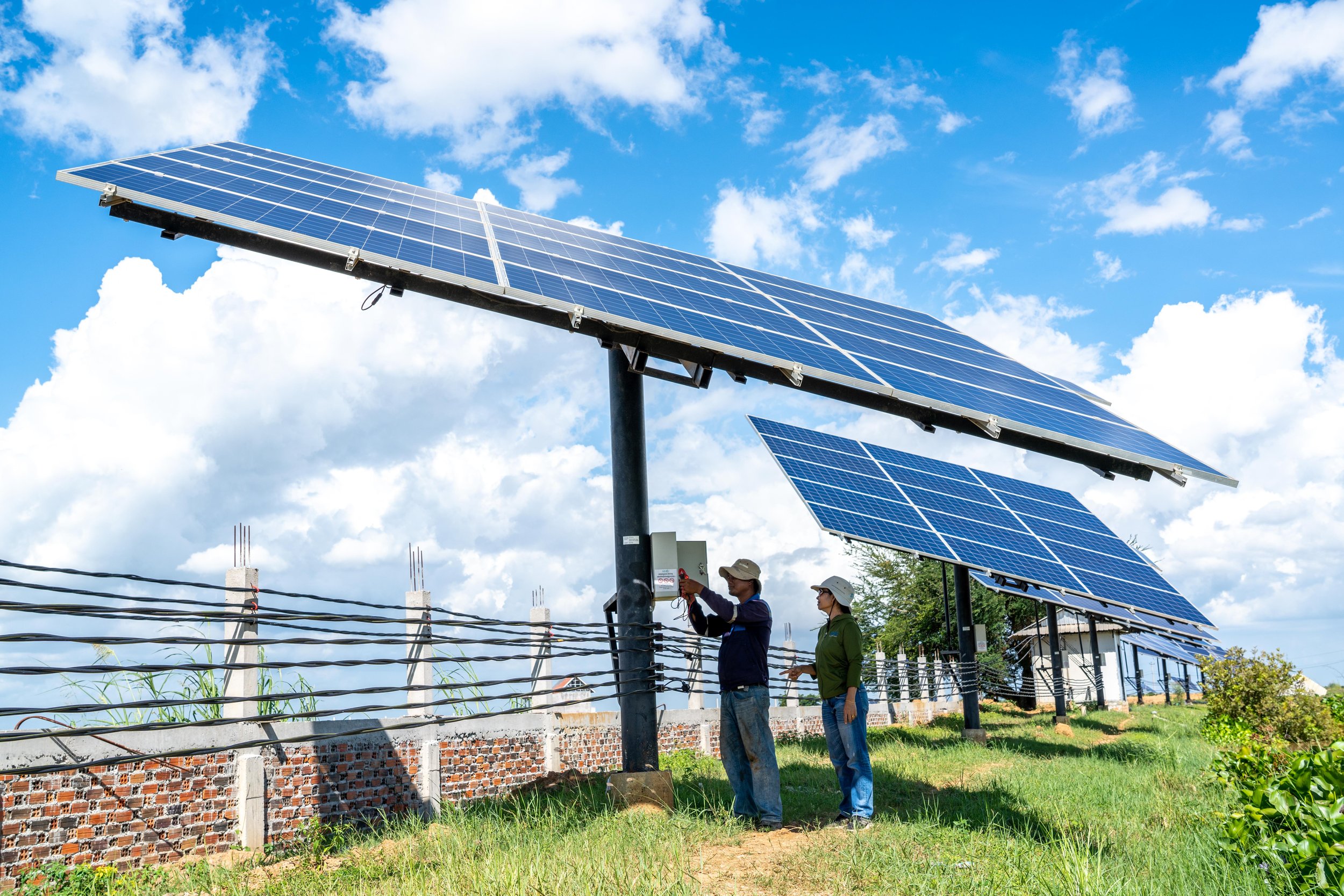 Two workers wearing helmets installing solar panels on a sunny day with a partly cloudy sky.