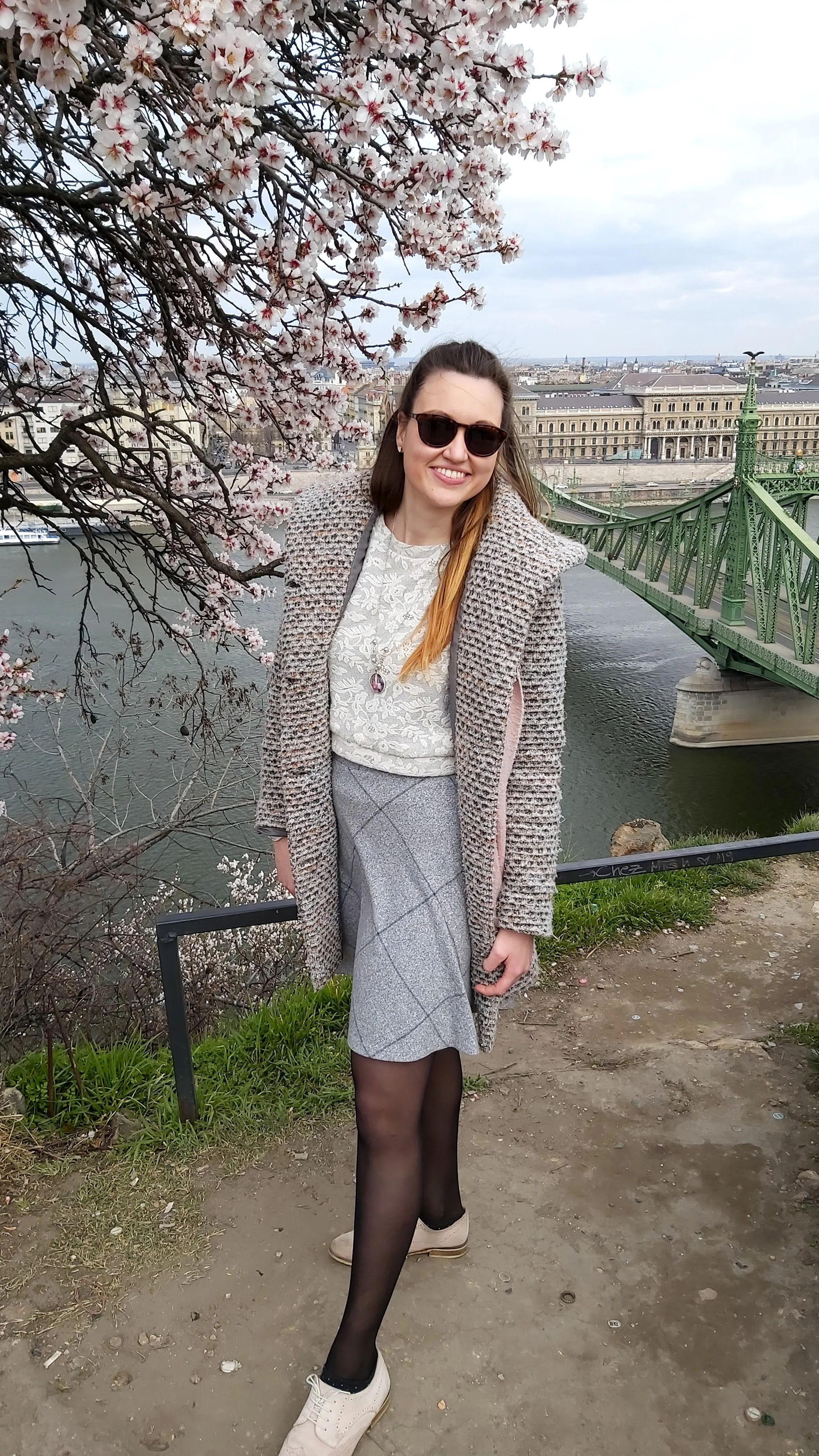 A woman in sunglasses smiling and standing by the river on a ledge, with cherry blossoms in full bloom and a bridge in the background.