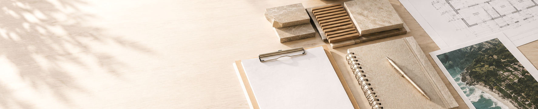 A desk with architectural plans, photographs, notebooks, and material samples, illuminated by natural light.