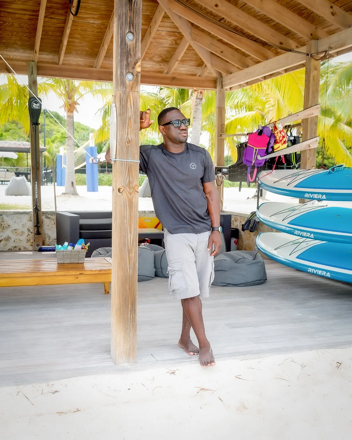 A Man in Sunglasses Leaning against a Pavilion Post Next to Stacked Riviera Paddle boards