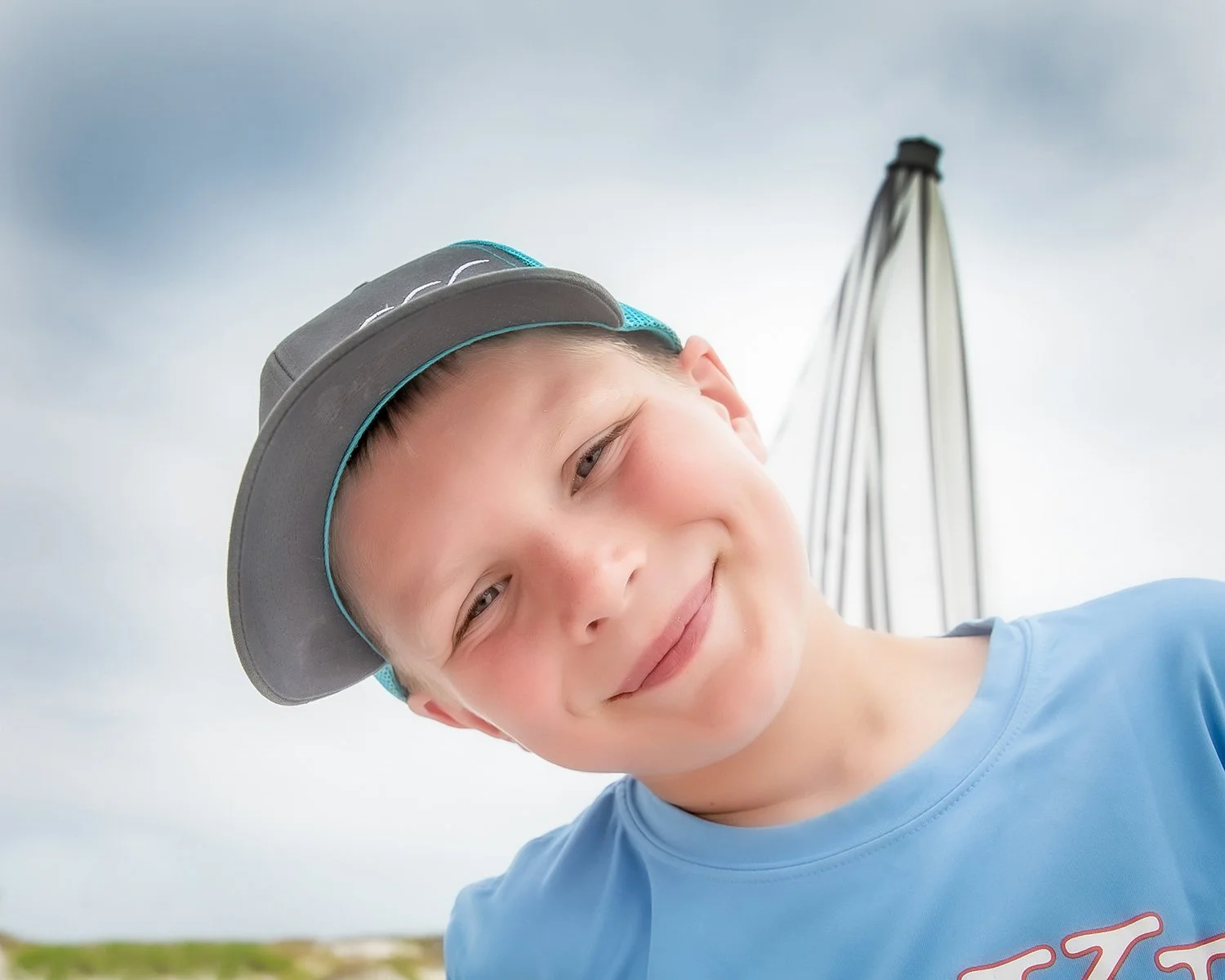 A Young Boy in a Baseball Cap Tilting his Head and Smiling under a Cloudy Sky