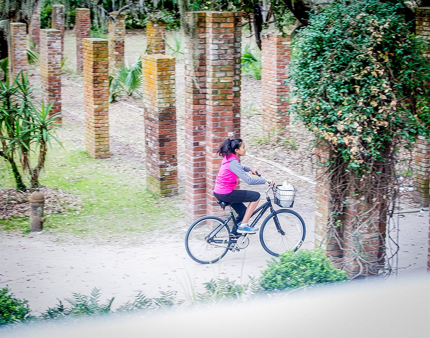 A Young Woman in a Pink Sweatshirt Riding a Bicycle Down a Path Lined with Brick Pillars
