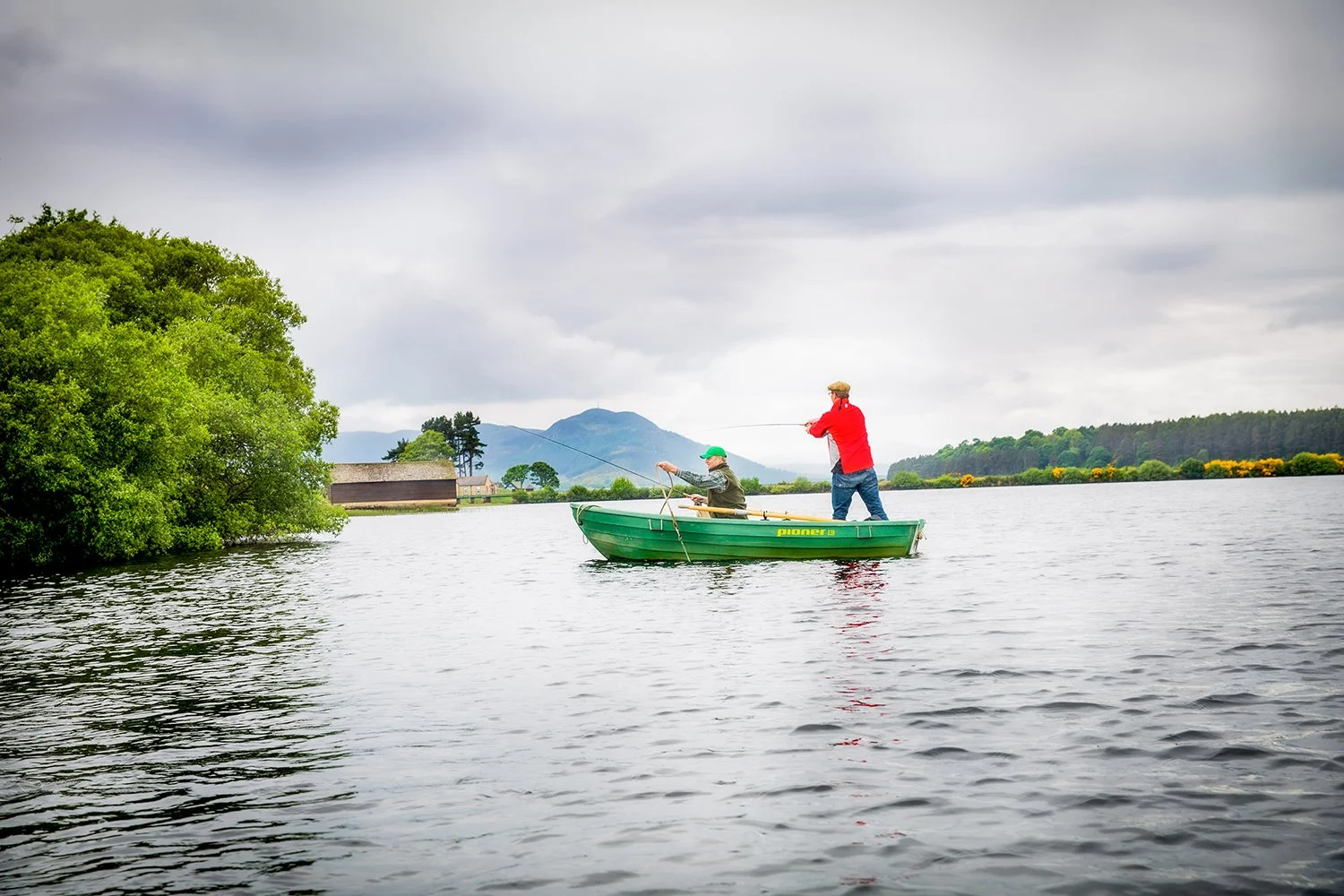 Two Men Fly Fishing from a Green Pioneer Rowboat on a Scenic River with Mountains Behind