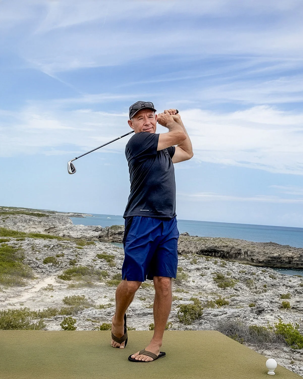 On-Location Lifestyle Portrait of a Senior Man in Baseball Cap and Flip-Flops Golfing on a Cliffside Tee Box Overlooking the Ocean