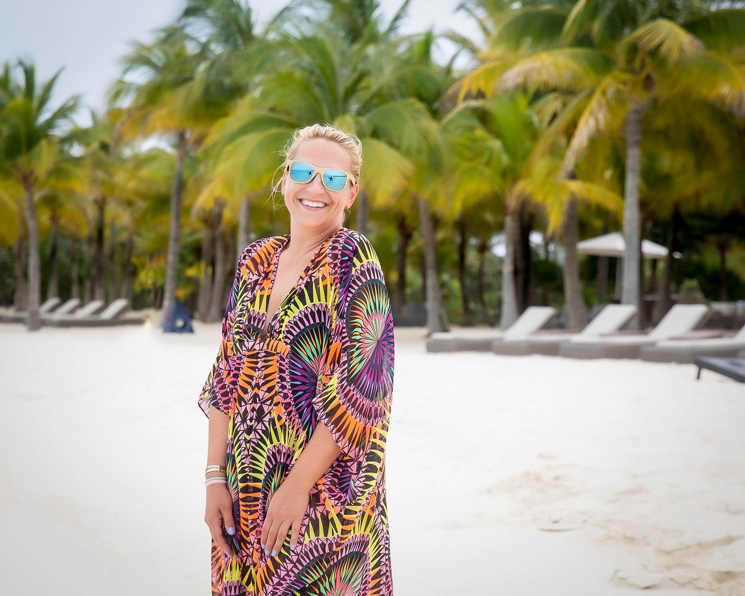 A Smiling Woman in Reflective Blue Sunglasses and a Colorful Patterned Kaftan on a Beach Lined with Palm Trees