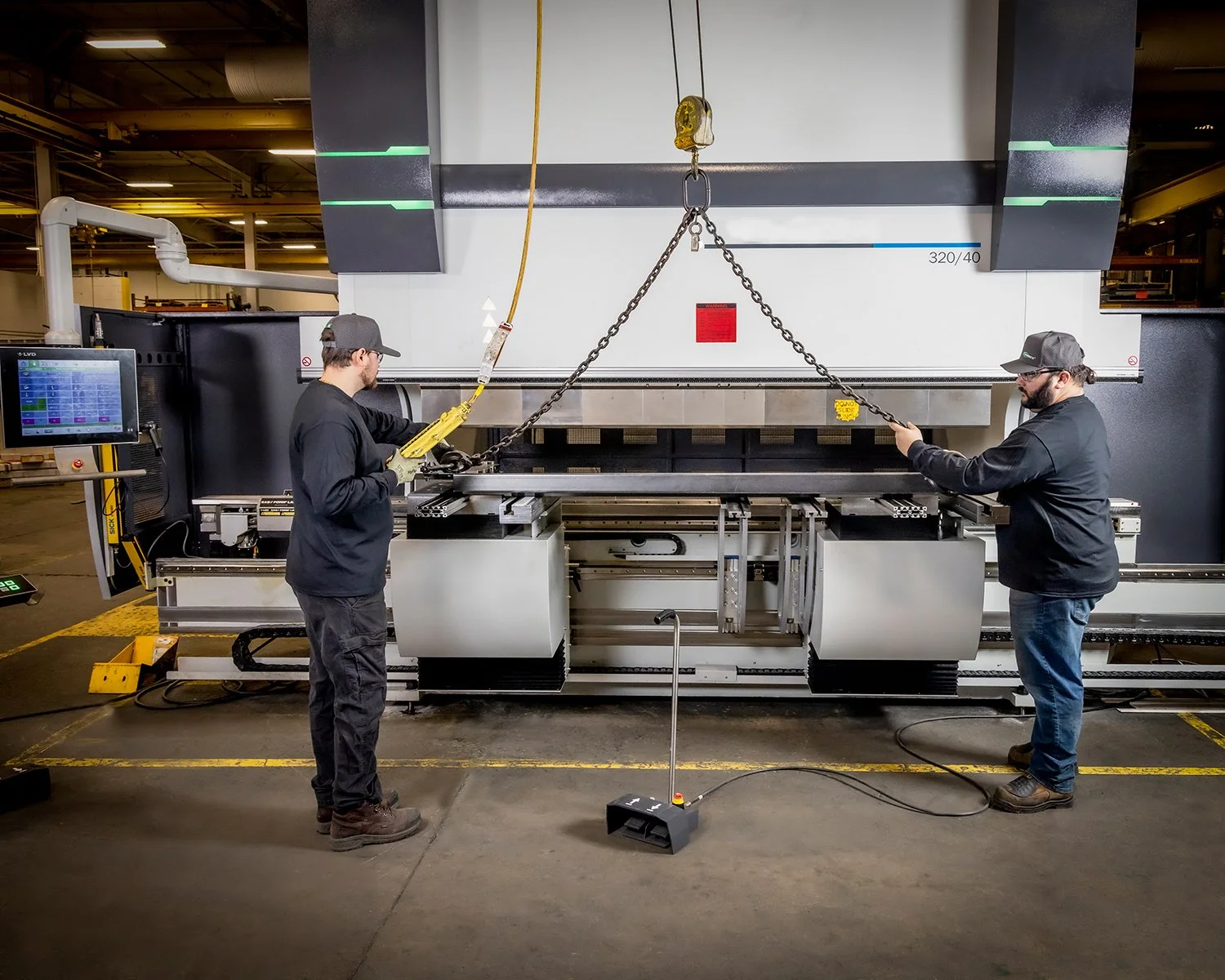 Industrial Factory Photography of Factory Technicians Using an Overhead Hoist Crane to Position Metal Sheets Inside an Industrial CNC Press Brake Machine