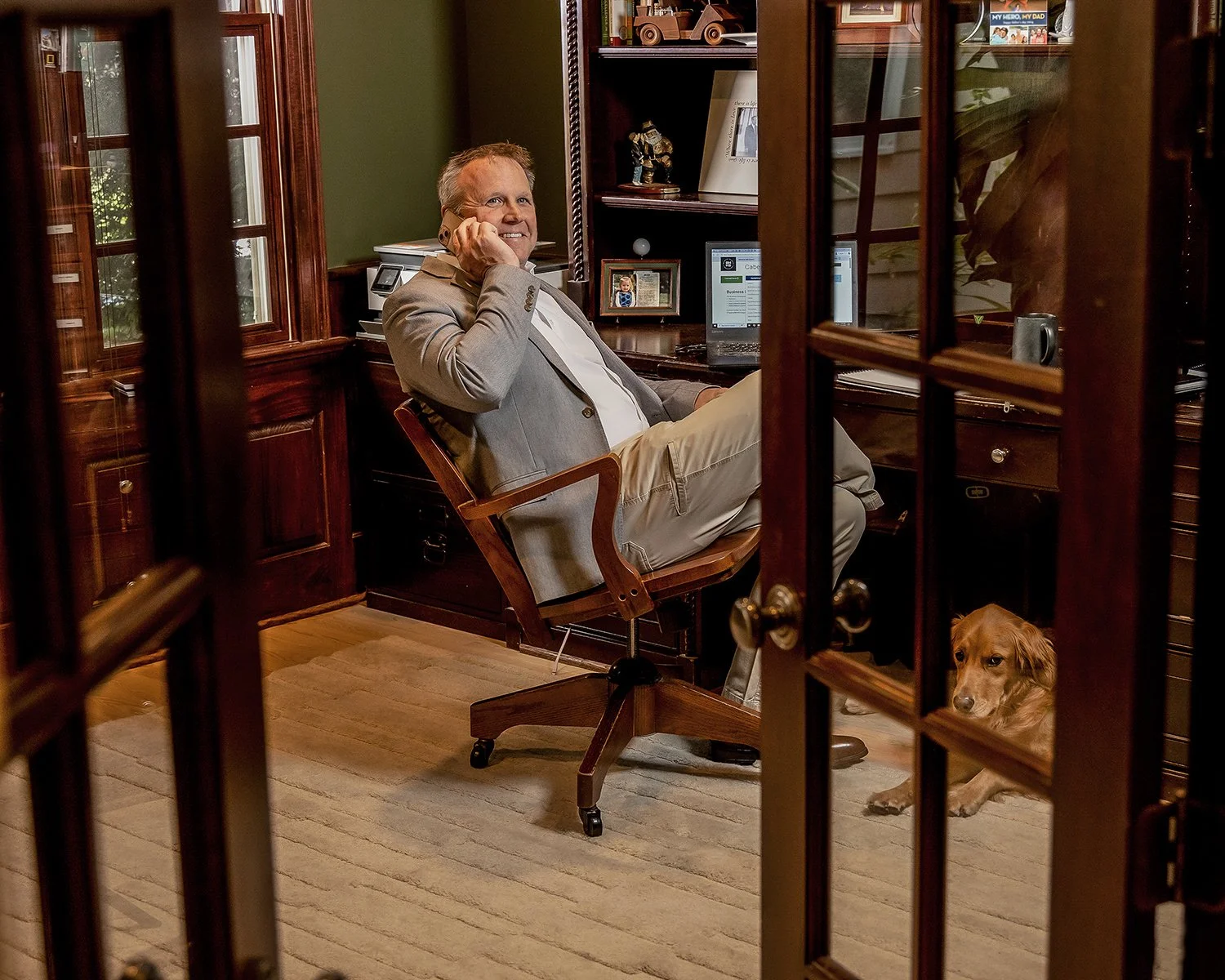 Financial Executive Talking on the Phone in a Wood-Paneled Home Office with a Golden Retriever Viewed Through French Doors