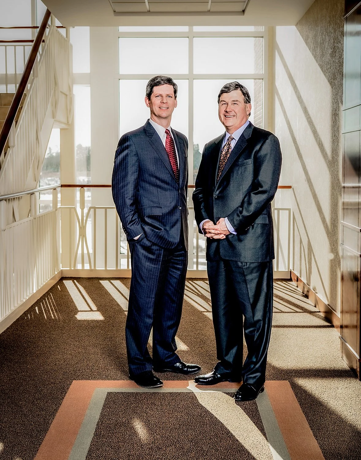 Two Executives Wearing Dark Suits in a Bright Hallway in Front of a Large Window 