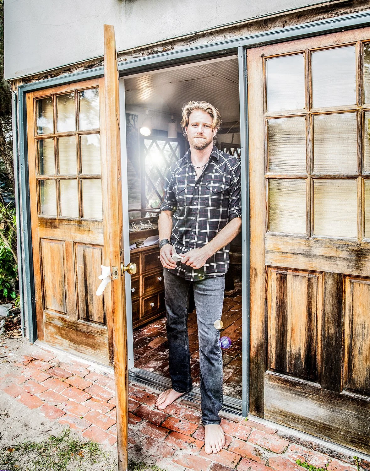 Commercial Lifestyle Portrait of a Blonde Man Standing Barefoot by a Doorway of a Rustic Workshop