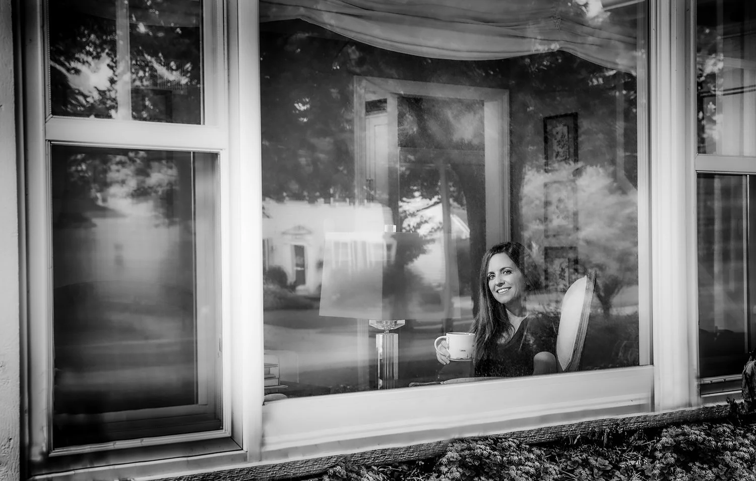 Black and White Portrait of a Woman Sitting Down in a Living Room Holding a Coffee Mug Looking Through a Large Glass Window with Reflections