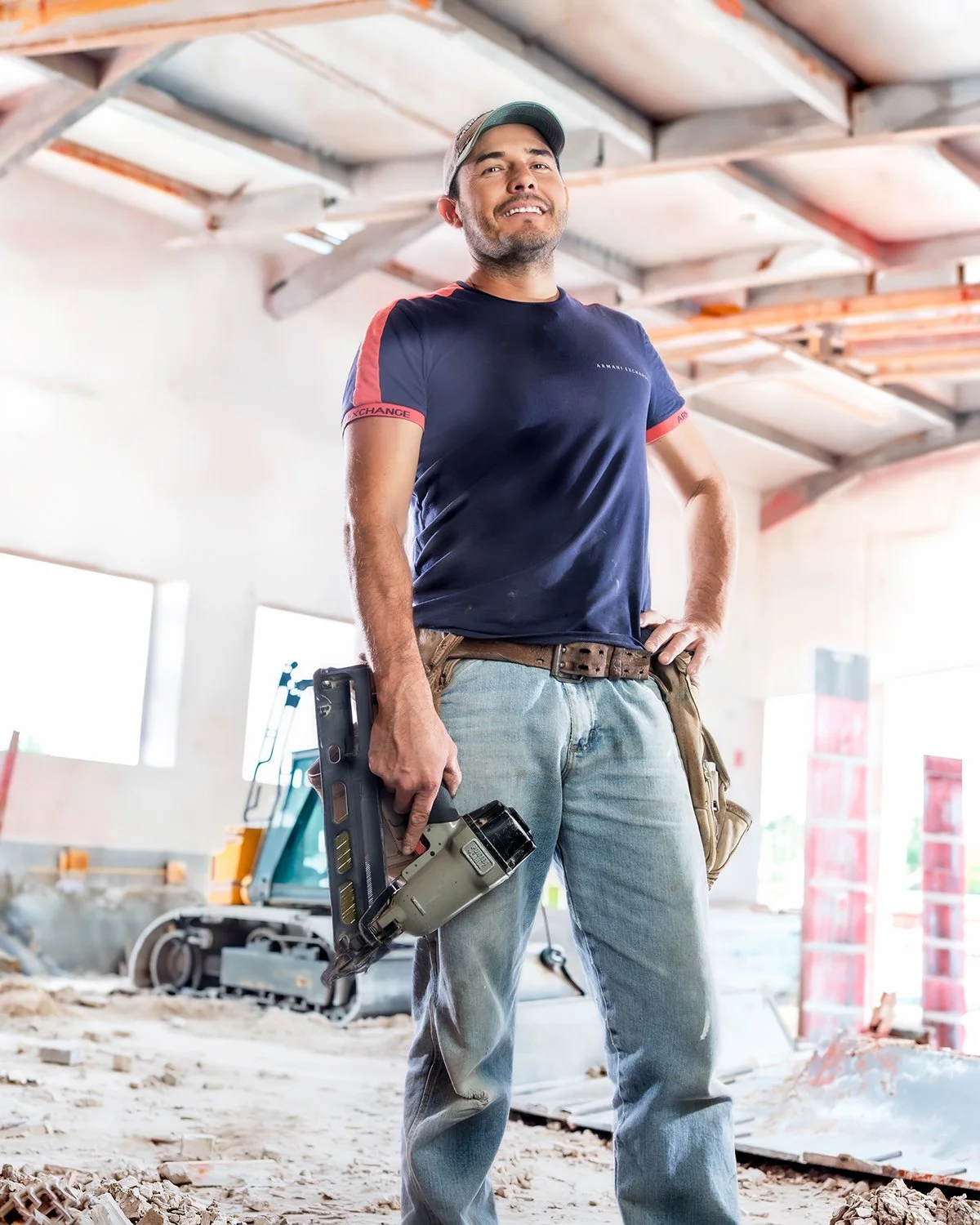 Smiling Male Contractor with a Tool Belt and Nail Gun Posing Inside an Under-Construction Industrial Building